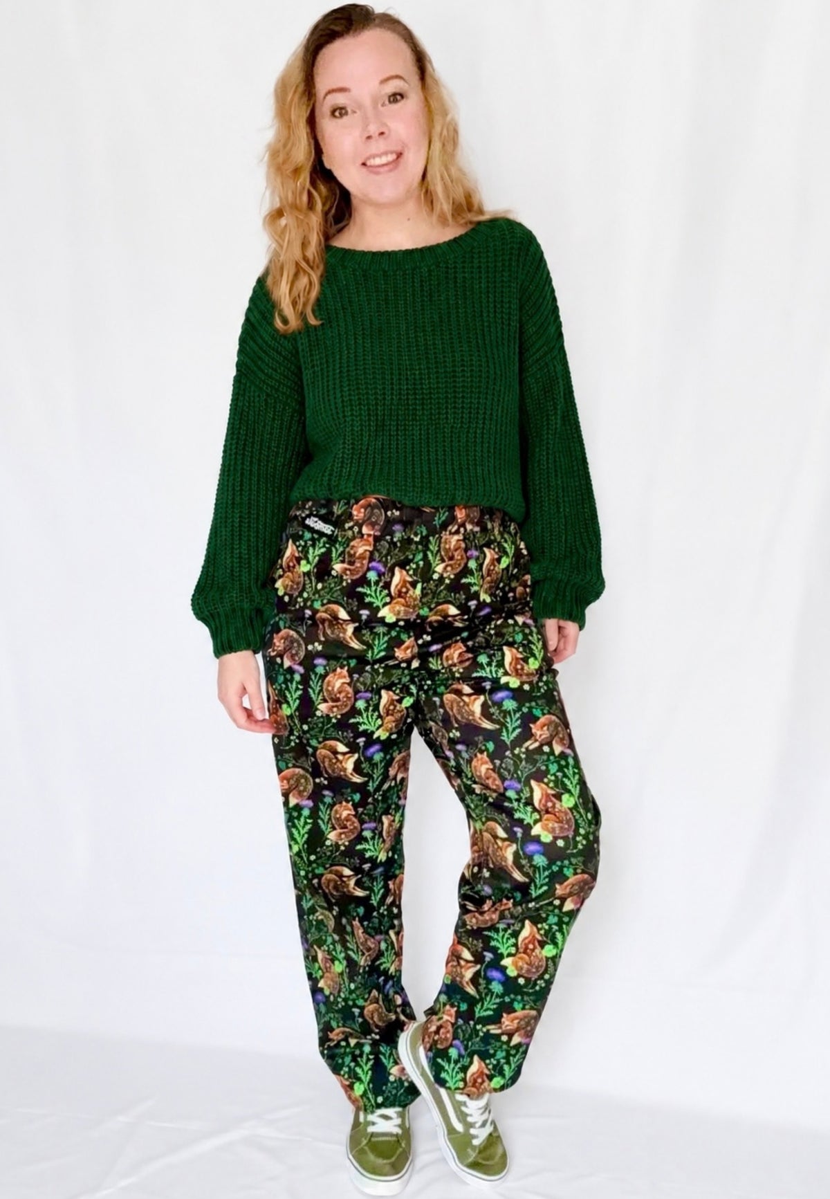 A woman modelling a dark green ribbed knit sweater and black trousers with a beautiful fox and thistle print, standing against a white studio background