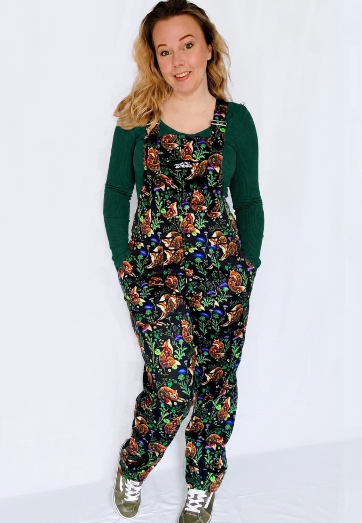 A woman modelling a pair of black dungarees with a beautiful, nature inspired print of fixes and thistles over a dark green long sleeve top, standing against a plain white studio background.