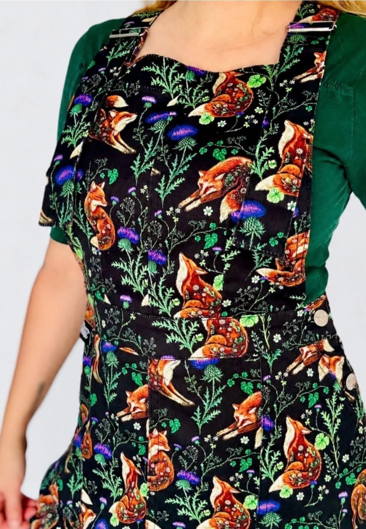 A close up of a person wearing a black pinafore dress with a colourful woodland print of foxes, green leaves, and purple thistles, layered over a dark green short sleeve top.