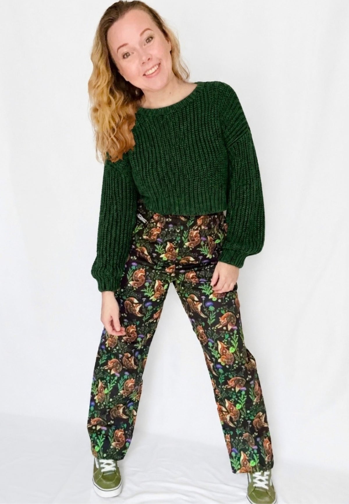 A woman modelling a dark green ribbed knit sweater and black trousers with a beautiful fox and thistle print, standing against a white studio background.