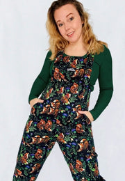A woman modelling a pair of black dungarees with a beautiful, nature inspired print of fixes and thistles over a dark green long sleeve top, standing against a plain white studio background.