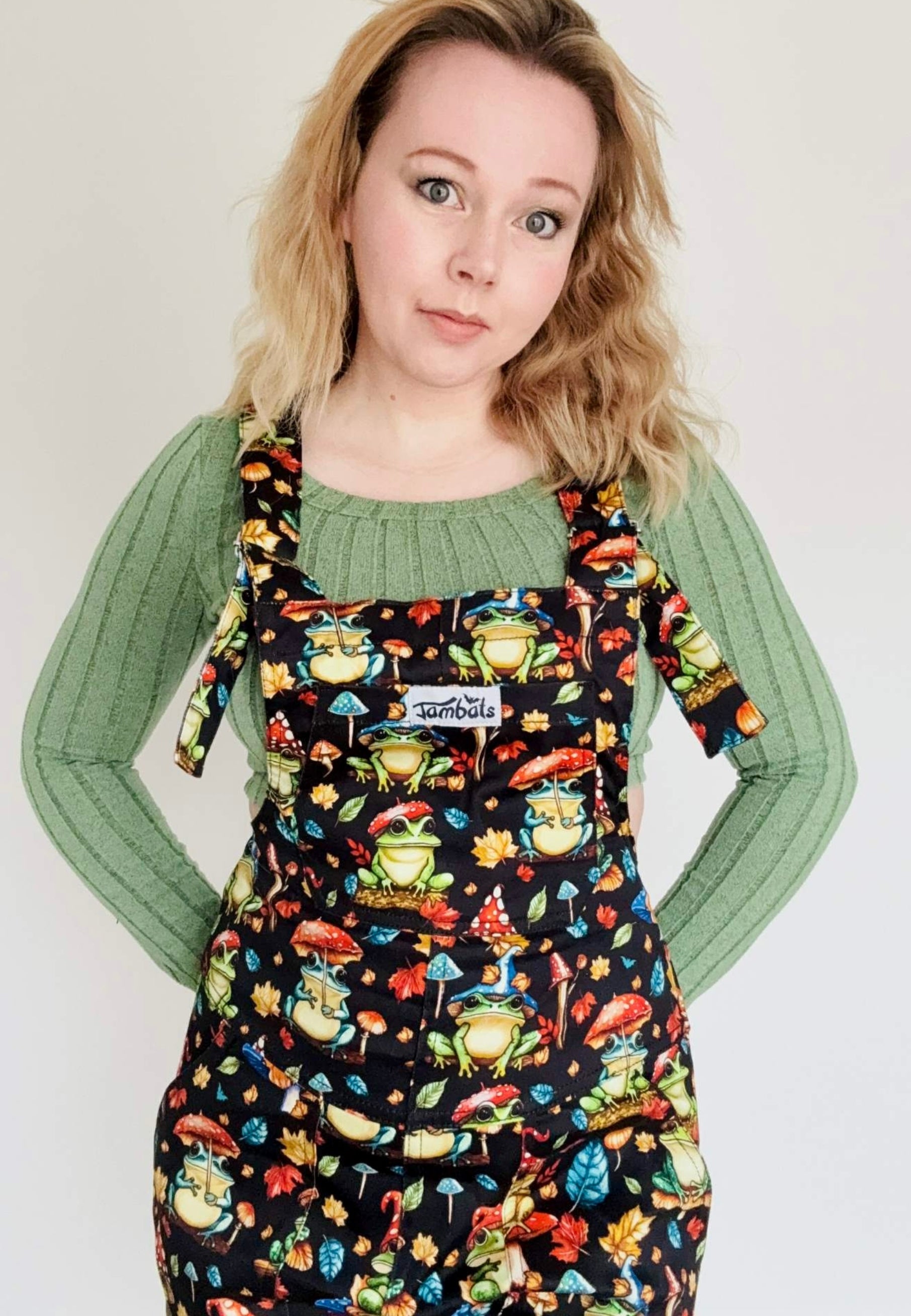Woman wearing dungarees with a whimsical print of forest frogs with toadstool hats and toadstool umbrellas. Studio photo. 