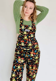 Woman wearing dungarees with a whimsical print of forest frogs with toadstool hats and toadstool umbrellas. Studio photo. 