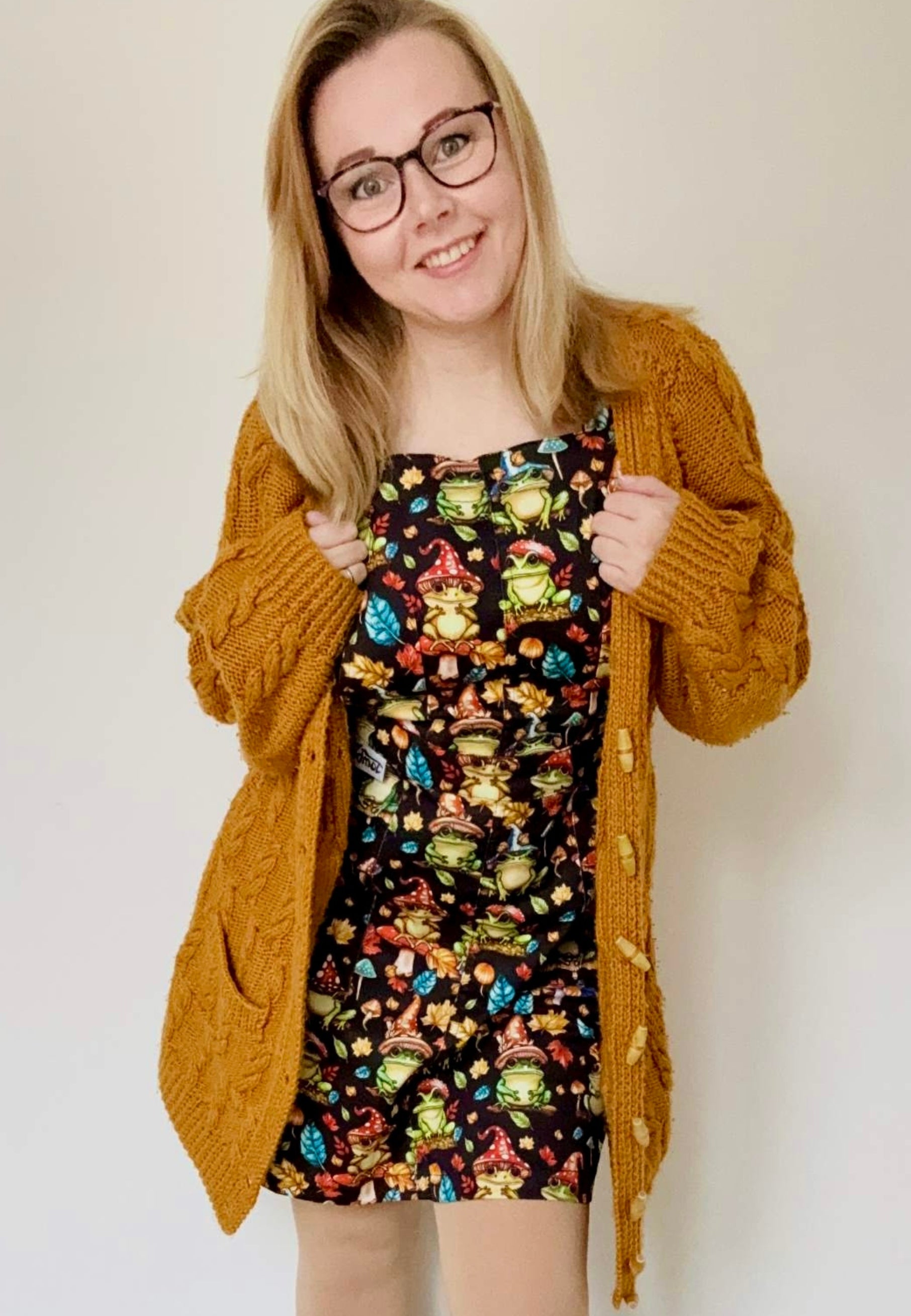 Woman in bookish glasses wearing a pinafore dress with a whimsical print of frogs with toadstool hats and toadstool umbrellas. Studio photo. 