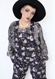 A model wearing spooky gothic dungarees with a purple and black cat ghost print, a grey ruffled cardigan, a black wide brim witch hat, and a chunky gold necklace against a plain white background.