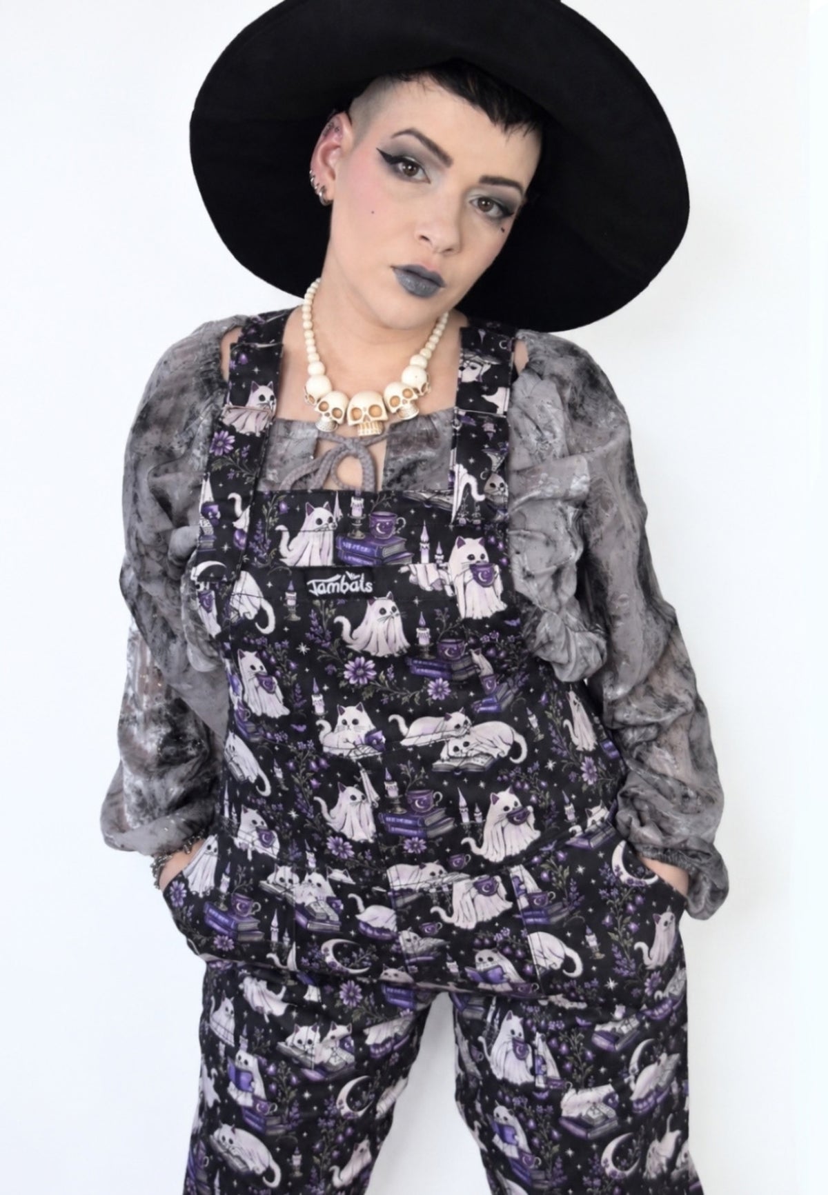 A model wearing spooky gothic dungarees with a purple and black cat ghost print, a grey ruffled cardigan, a black wide brim witch hat, and a chunky gold necklace against a plain white background.