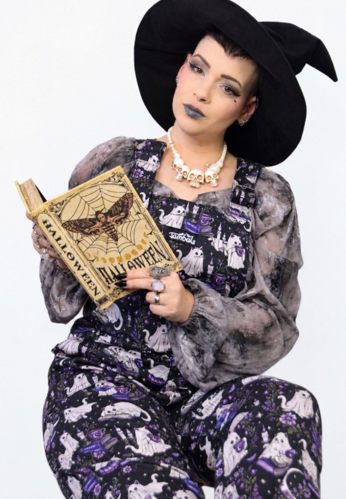 A woman wearing a black witch hat and black and purple dungarees with a ghost cats book club print, holding a vintage style Halloween book against a plain white background.