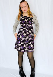 A woman modeling a black and purple ghost ca print pinafore dress over a light grey long sleeve polka dot top, paired with black tights and black lace up boots, against a white studio background.