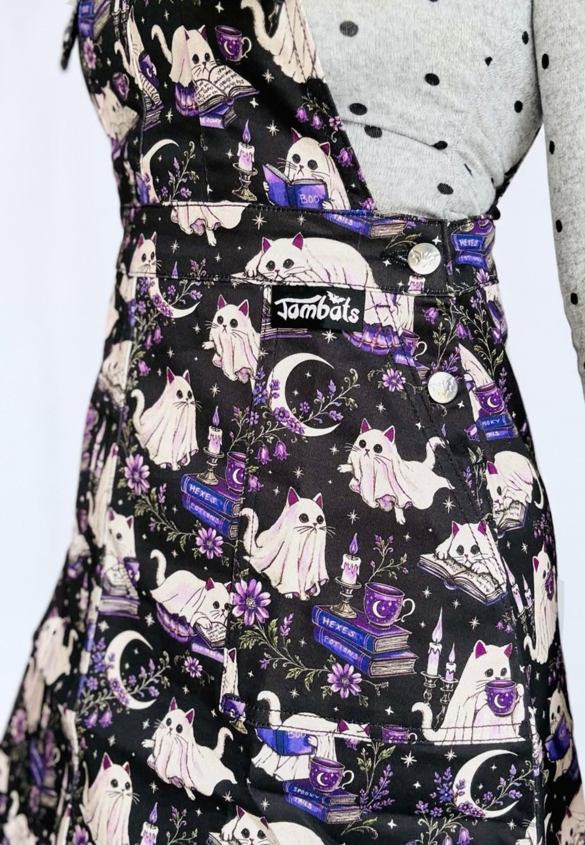 A close up of a person wearing a black pinafore dress with a spooky, whimsical print of white cats, purple flowers, candles, books, and moons. The garment has a visible small label on the waistband reading ‘Jambats' and is styled with a gray polka-dot top underneath.