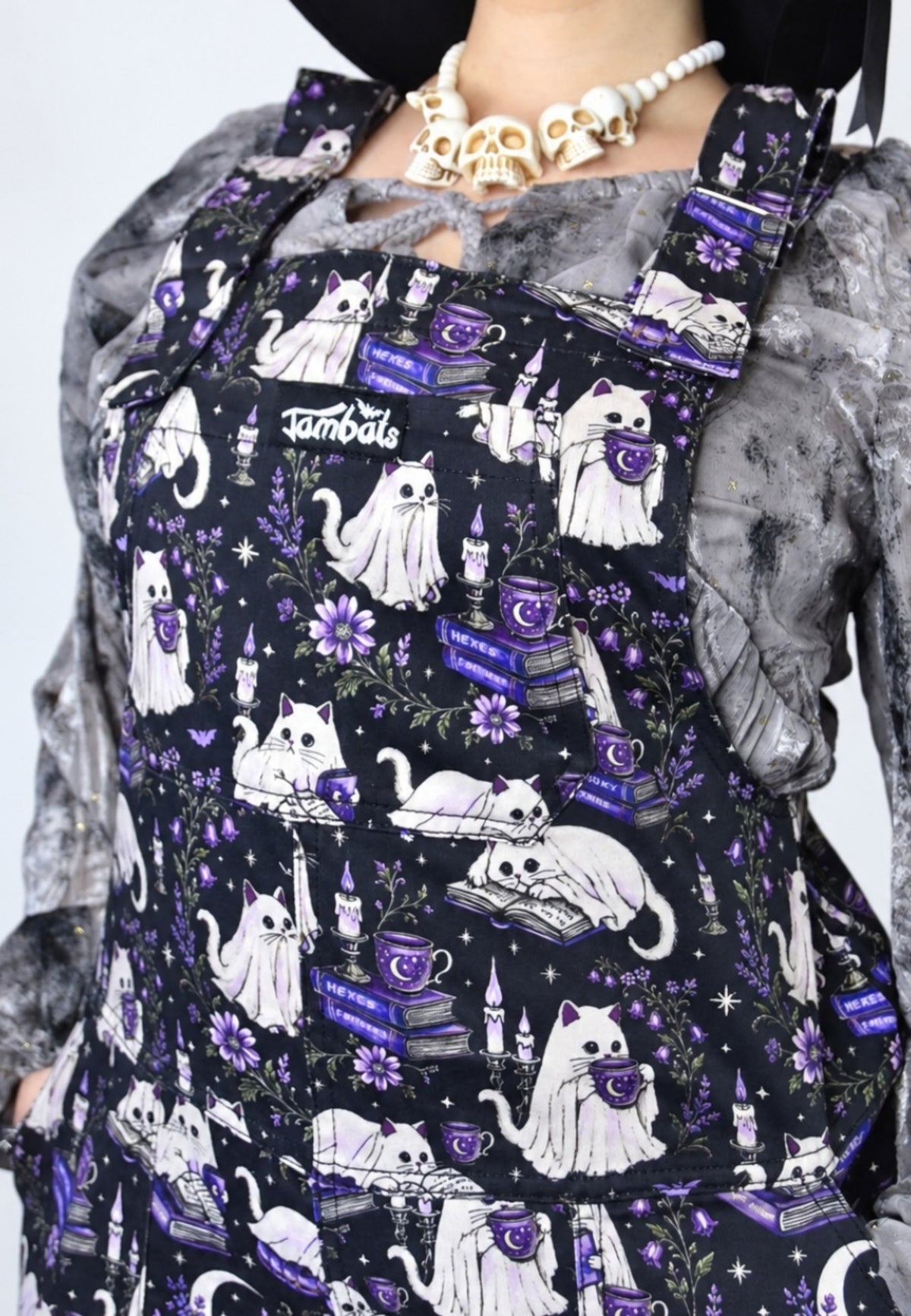 A close up of a person wearing black dungarees overalls with a repeat print of white ghost cats, purple books, candles and flowers, layered over a grey ruffled top and a gold skull necklace.