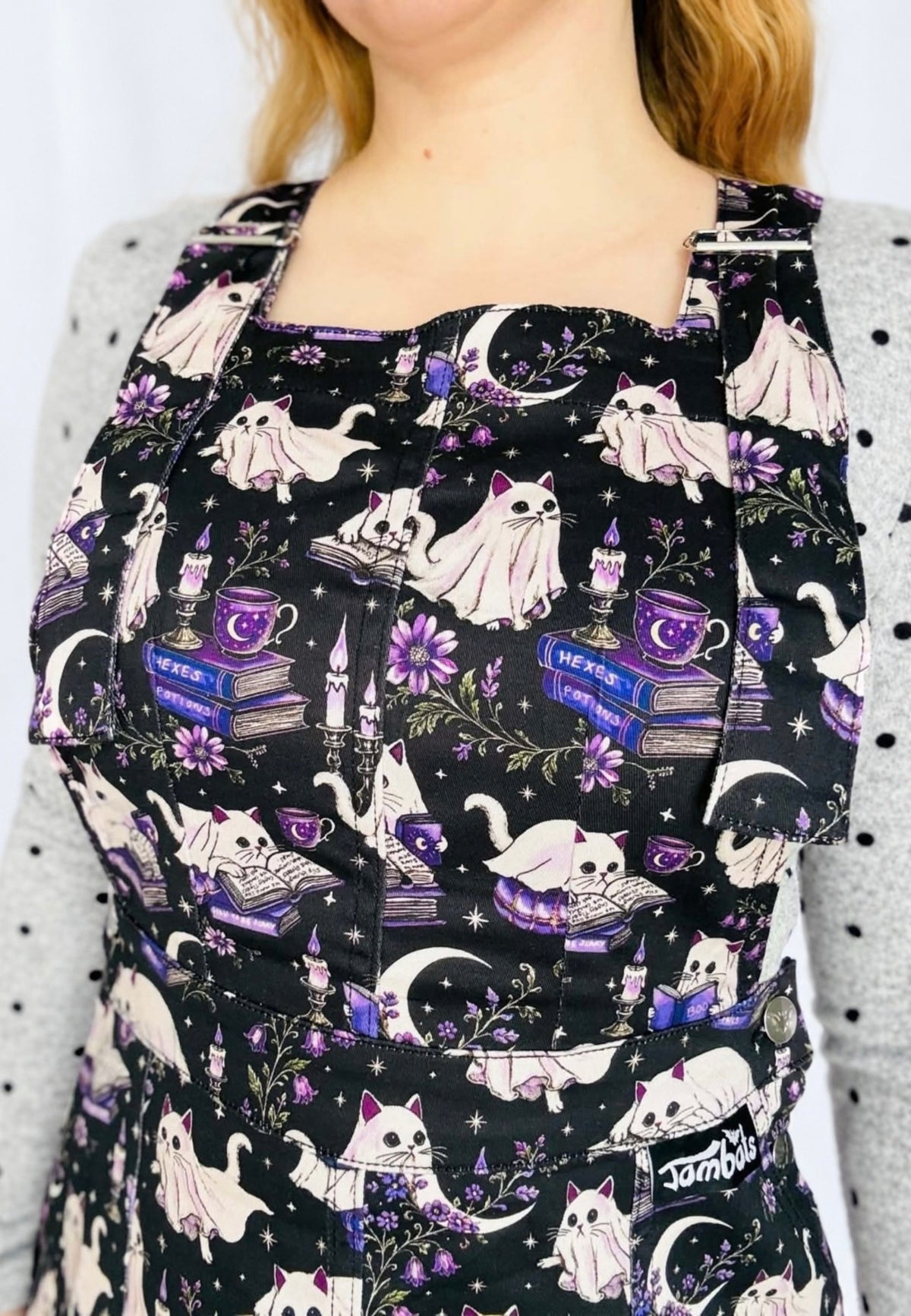 A close up of a person wearing a black pinafore dress with a spooky, whimsical print of white cats, purple flowers, candles, books, and moons. The garment is styled with a grey polka dot top underneath. 