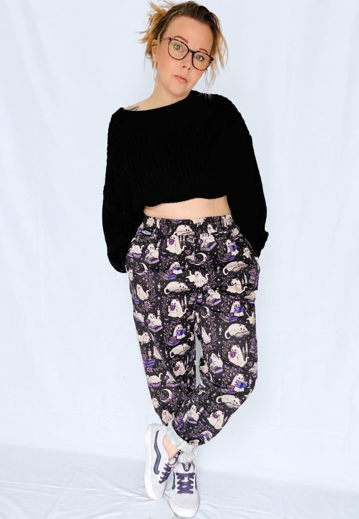 A woman modeling a black cropped knit sweater and black trousers with a spooky ghost cats print with white sneakers, standing against a plain light background.