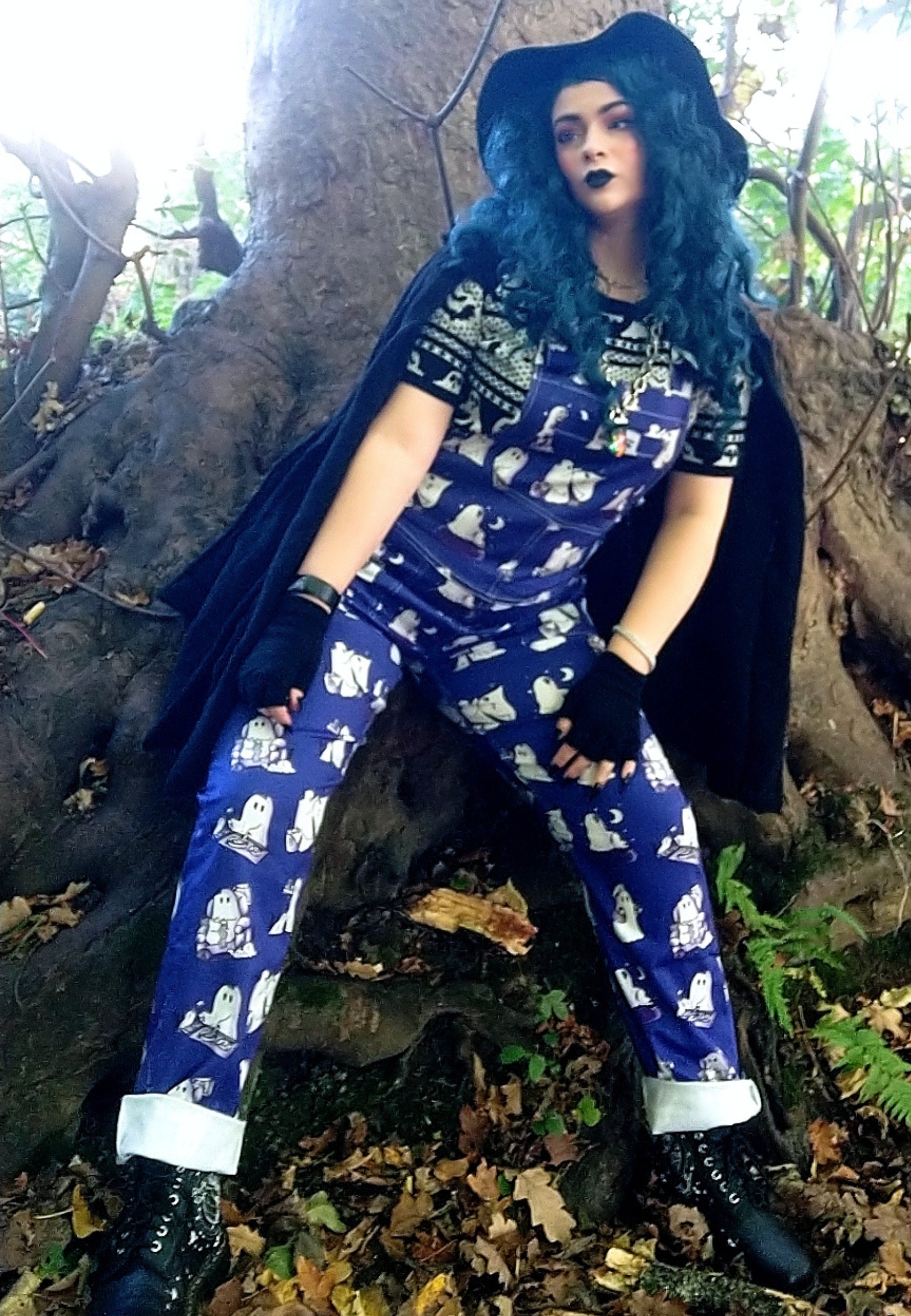A witchy woman in a witches hat and cloak outdoors in the woods  wearing cute blue dungarees with a print of ghosts and ghost cats. 
