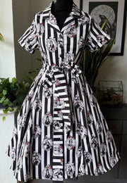 A short sleeve collared dress displayed on a black mannequin in an indoor room. The dress has bold gothic black and white vertical stripes with repeated circular vintage style portrait style pictures showing all the old school gothic ghouls. 