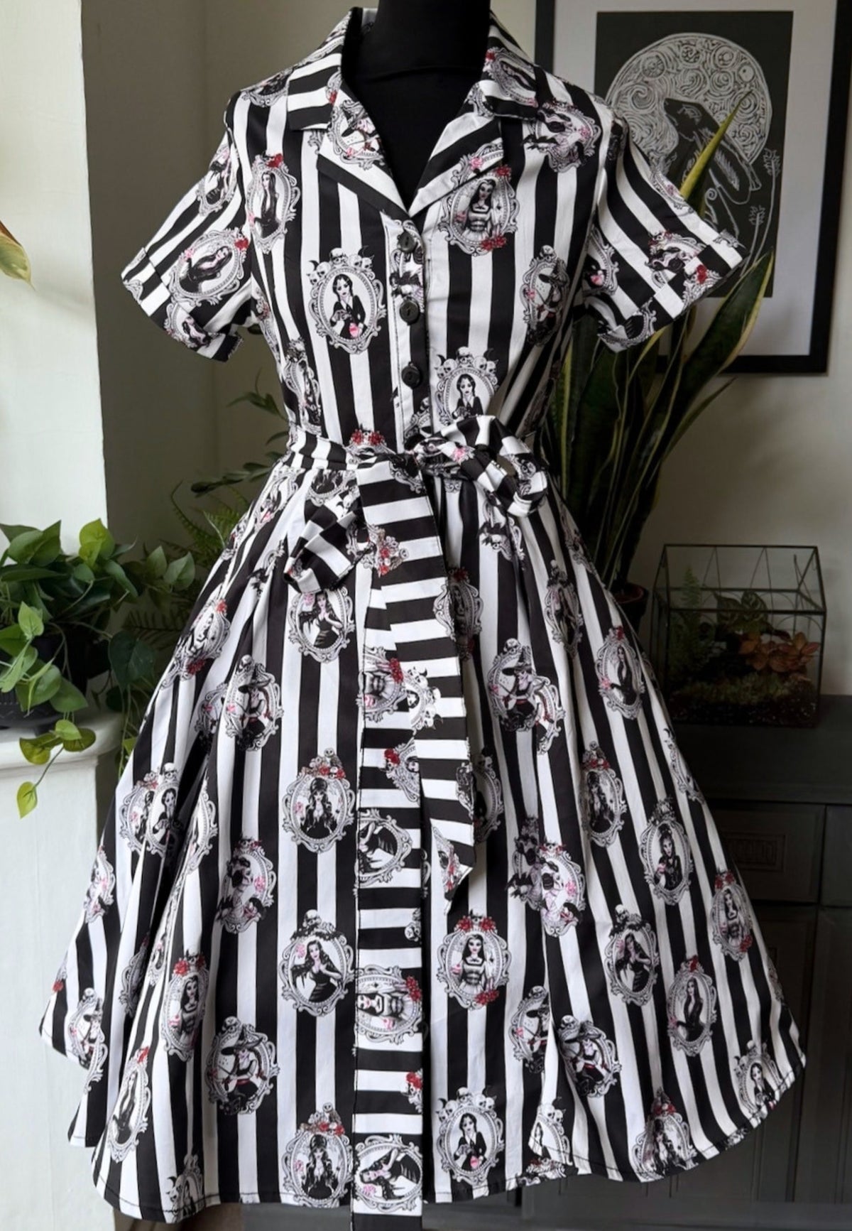 A short sleeve collared dress displayed on a black mannequin in an indoor room. The dress has bold gothic black and white vertical stripes with repeated circular vintage style portrait style pictures showing all the old school gothic ghouls. 