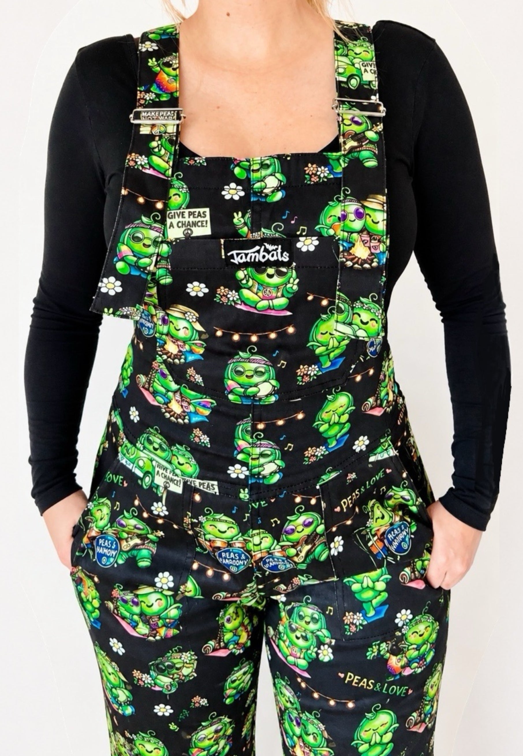 Woman wearing a black long-sleeve shirt and black dungarees overalls with a pattern of cute hippy peas at a music festival of pease and love. Close up of pattern. Woman is standing against a white background. 