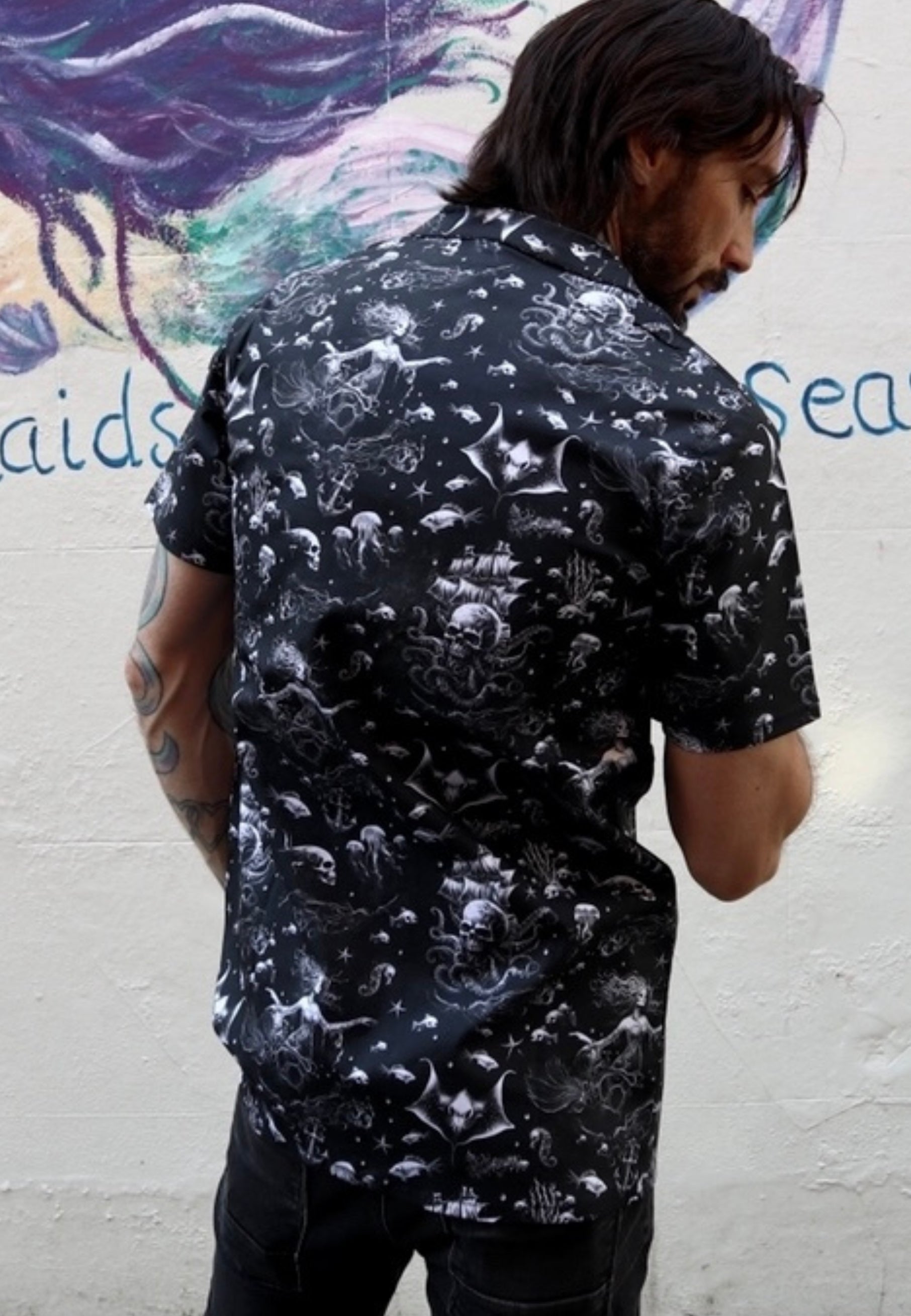 Back view of a man wearing a black shirt with a gothic under the sea print..ghostly mermaids and pirate ships. Man is standing in front of a mermaid wall mural.