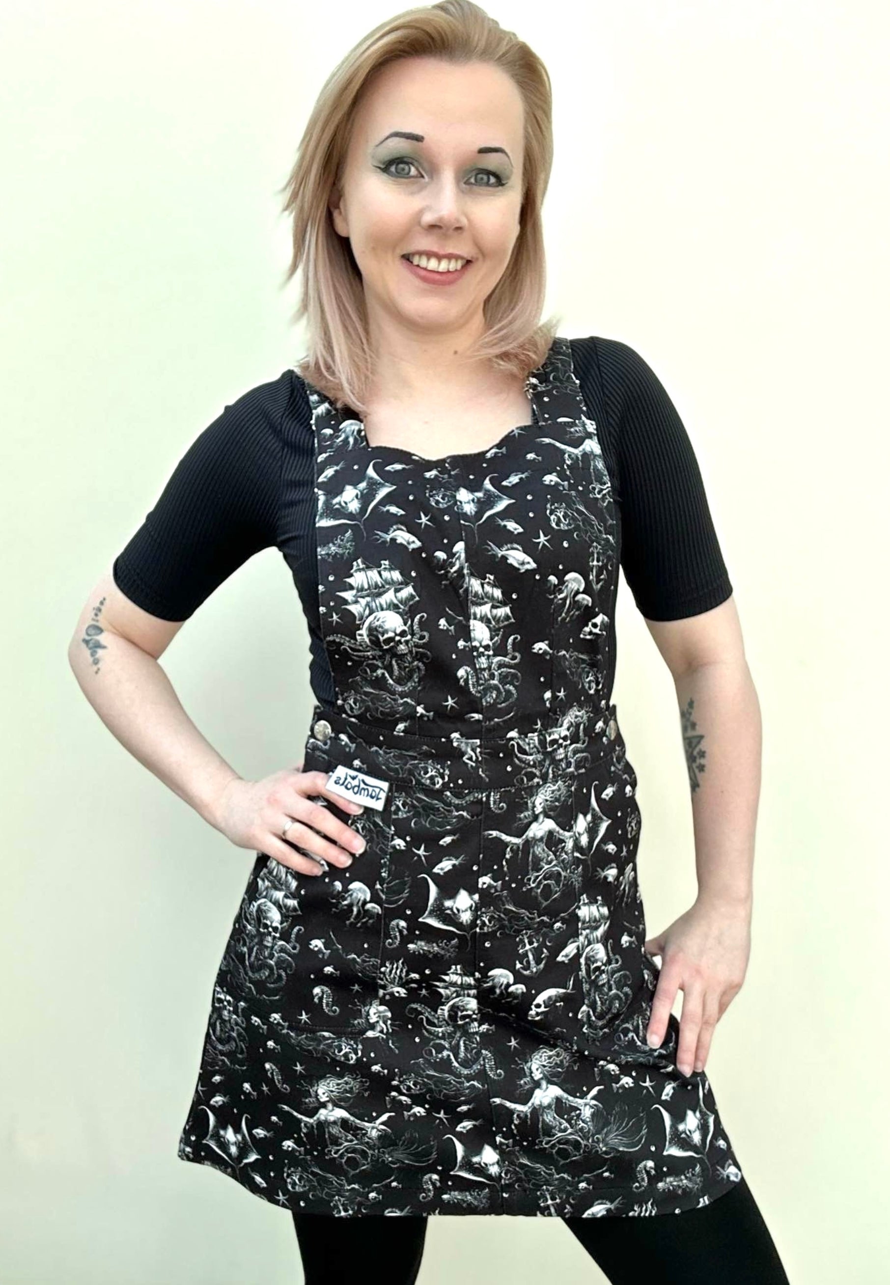 A woman wearing a black pinafore dress with a gothic underwater print, featuring deep hip pockets and side button fastenings. 