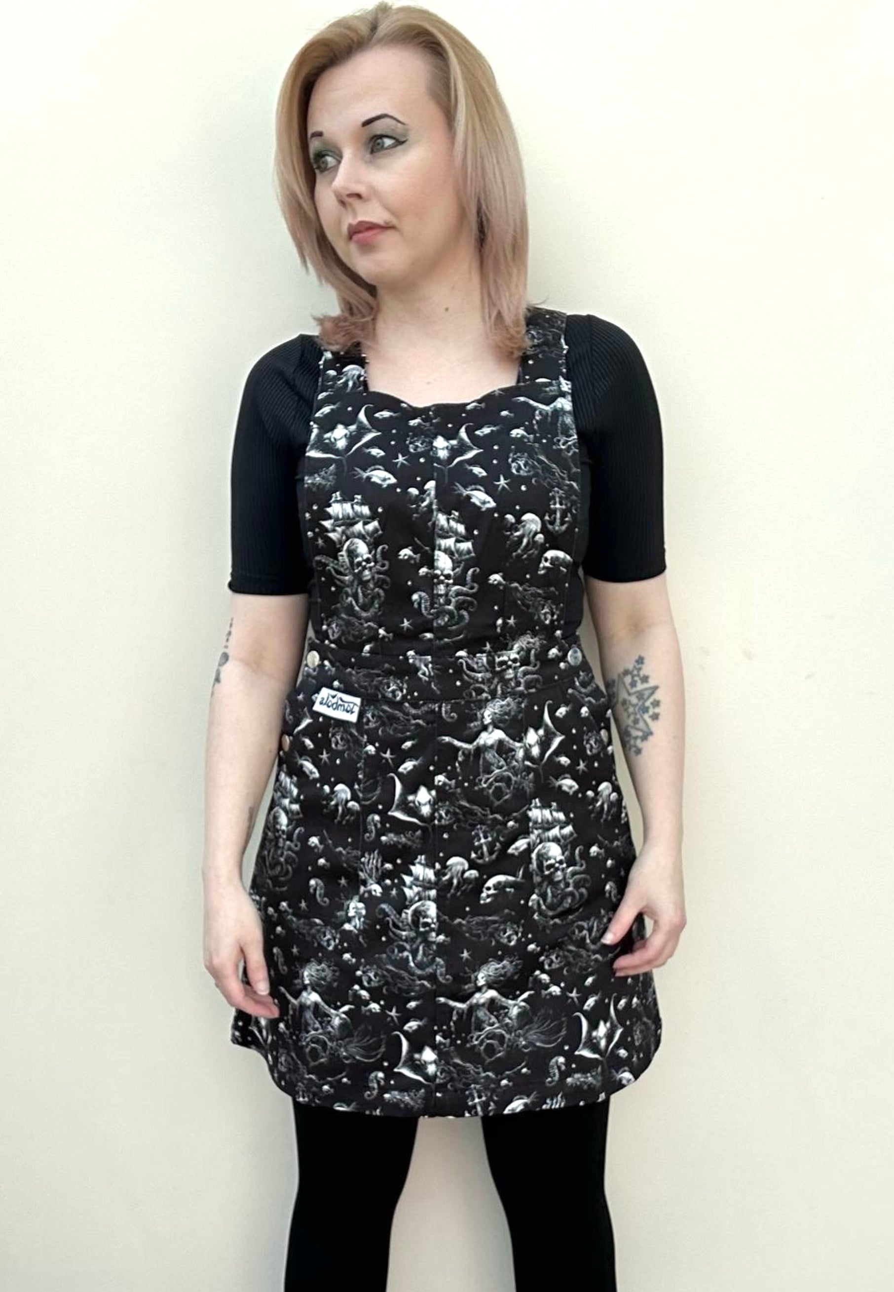 A person wearing a black pinafore dress with a gothic underwater print, featuring deep hip pockets and side button fastenings. 