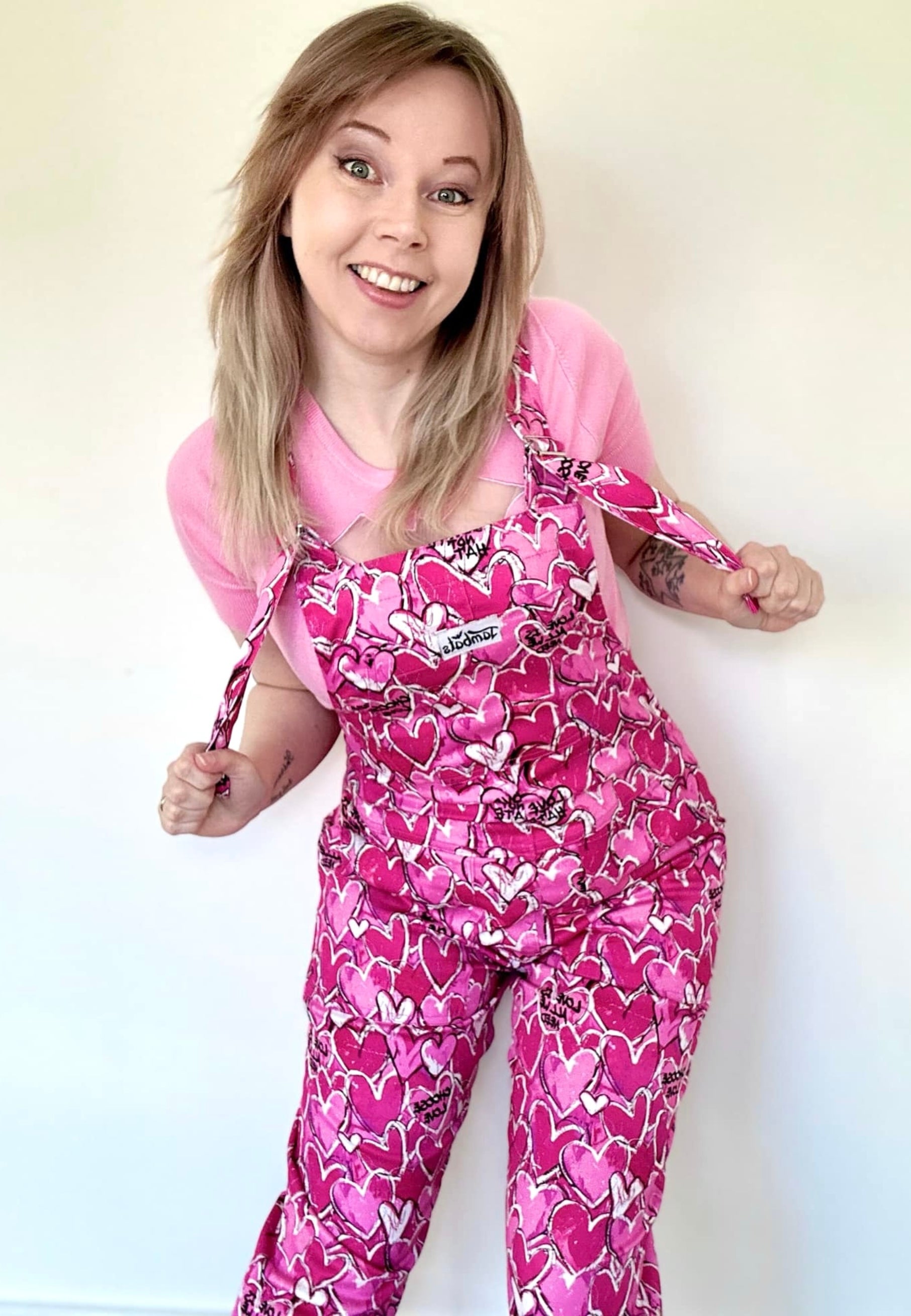 A woman wearing a pink and white heart patterned dungarees with a pink top, standing against a white background.