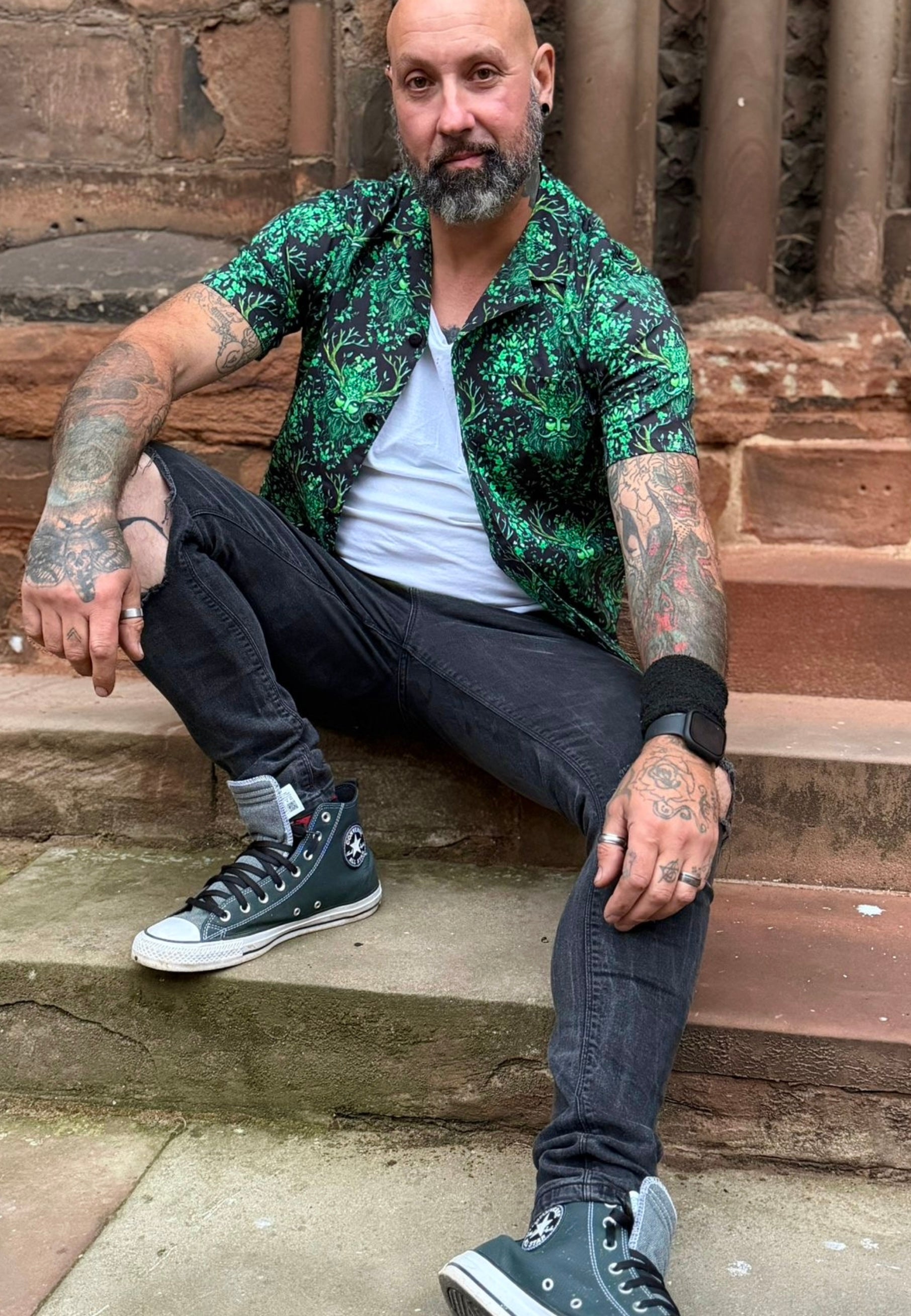 Man with tattoos is wearing a shirt with a Green Man print. Man is outdoors, sat on cathedral steps. 