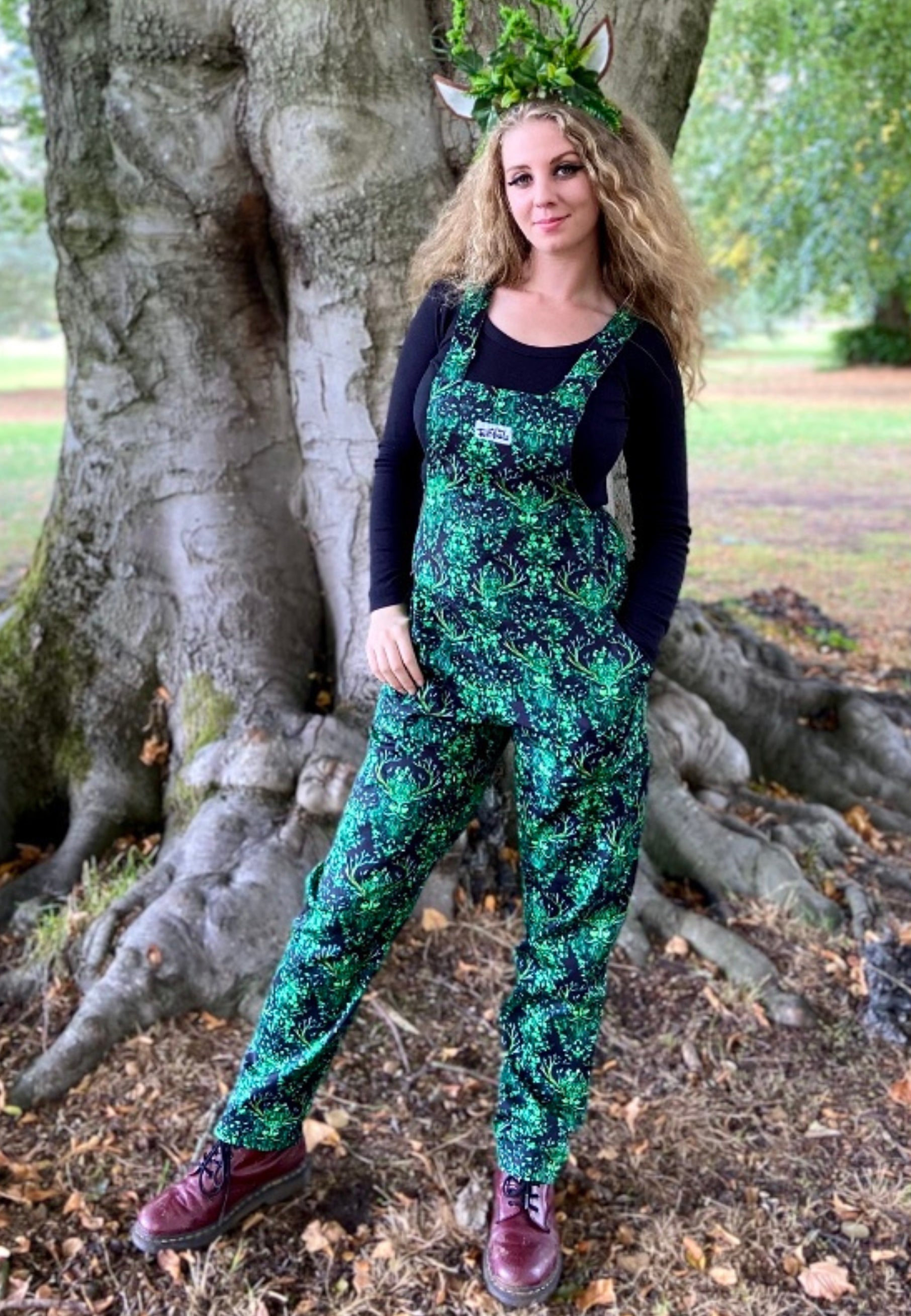 Woman standing in a forest setting wearing black dungarees with a beautiful green man print on the dungarees. Witchy pagan style.
