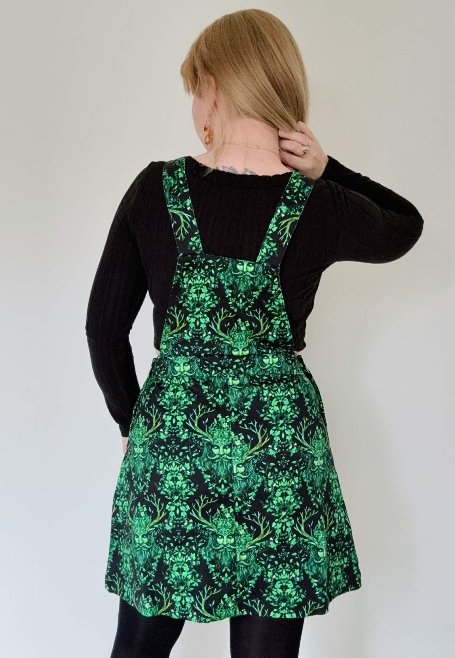 Back view of a woman wearing a black pinafore dress with a beautiful green print of the mythical green man. Studio photo on a plain white background. Witchy pagan style clothing aesthetic.