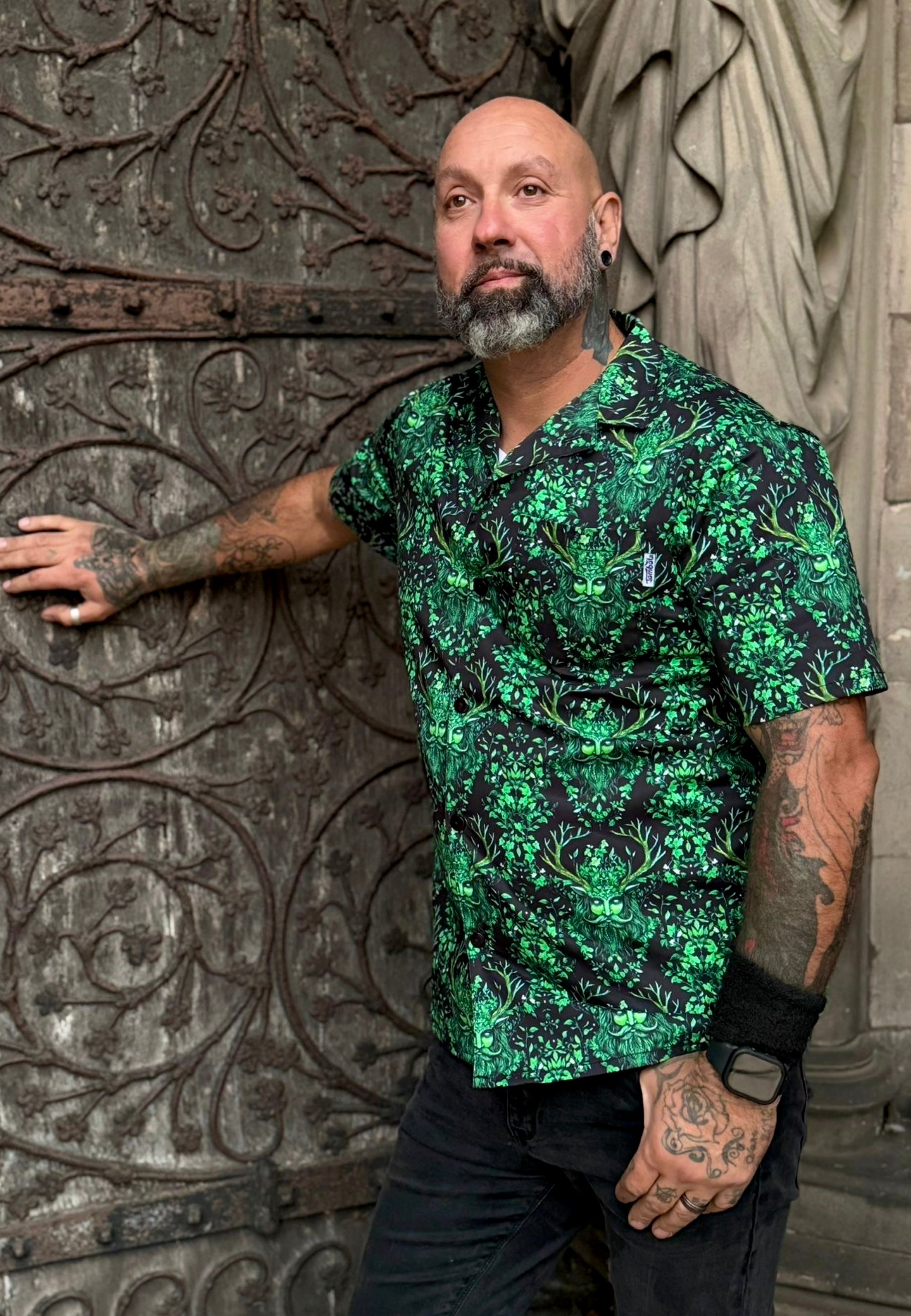 Man with tattoos is wearing a shirt with a Green Man print. Man is leaning against a big vintage cathedral door. 