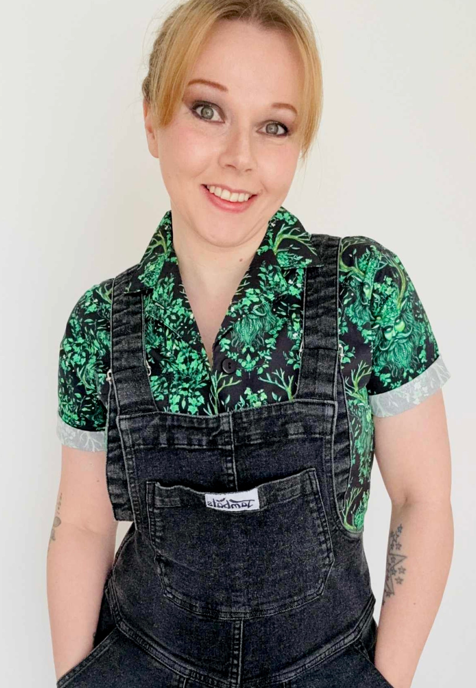 A woman wearing a Green Man print shirt and black dungarees. Studio photo. 