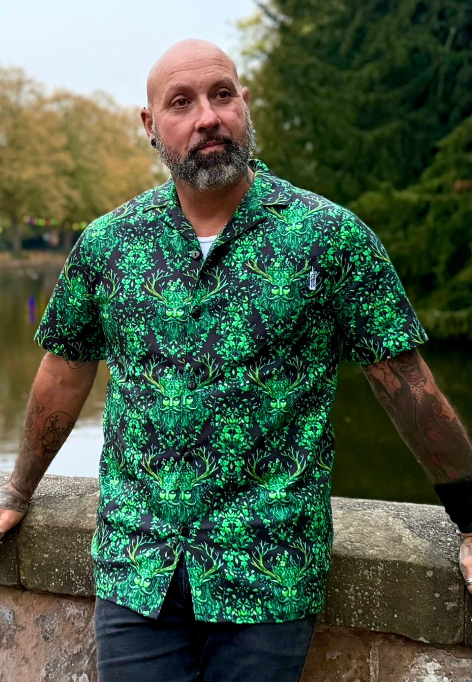 Man with tattoos is wearing a shirt with a Green Man print. Man is outdoors, natural pose. 