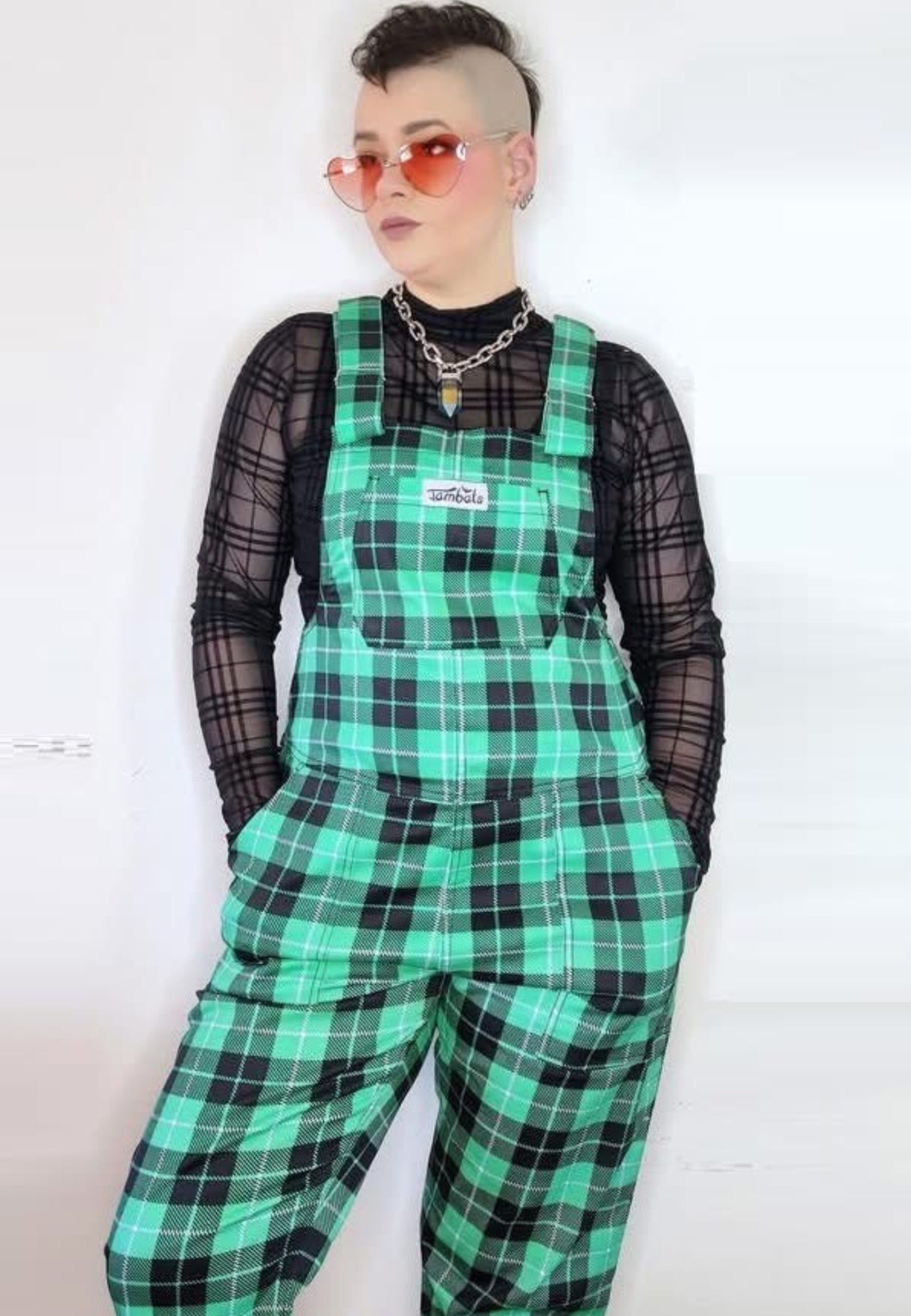 Model wearing green tartan dungarees, paired with a black mesh top and alternative sunglasses. The dungarees have multiple pockets and the person has their hands in the pockets. Alternative punk style. 