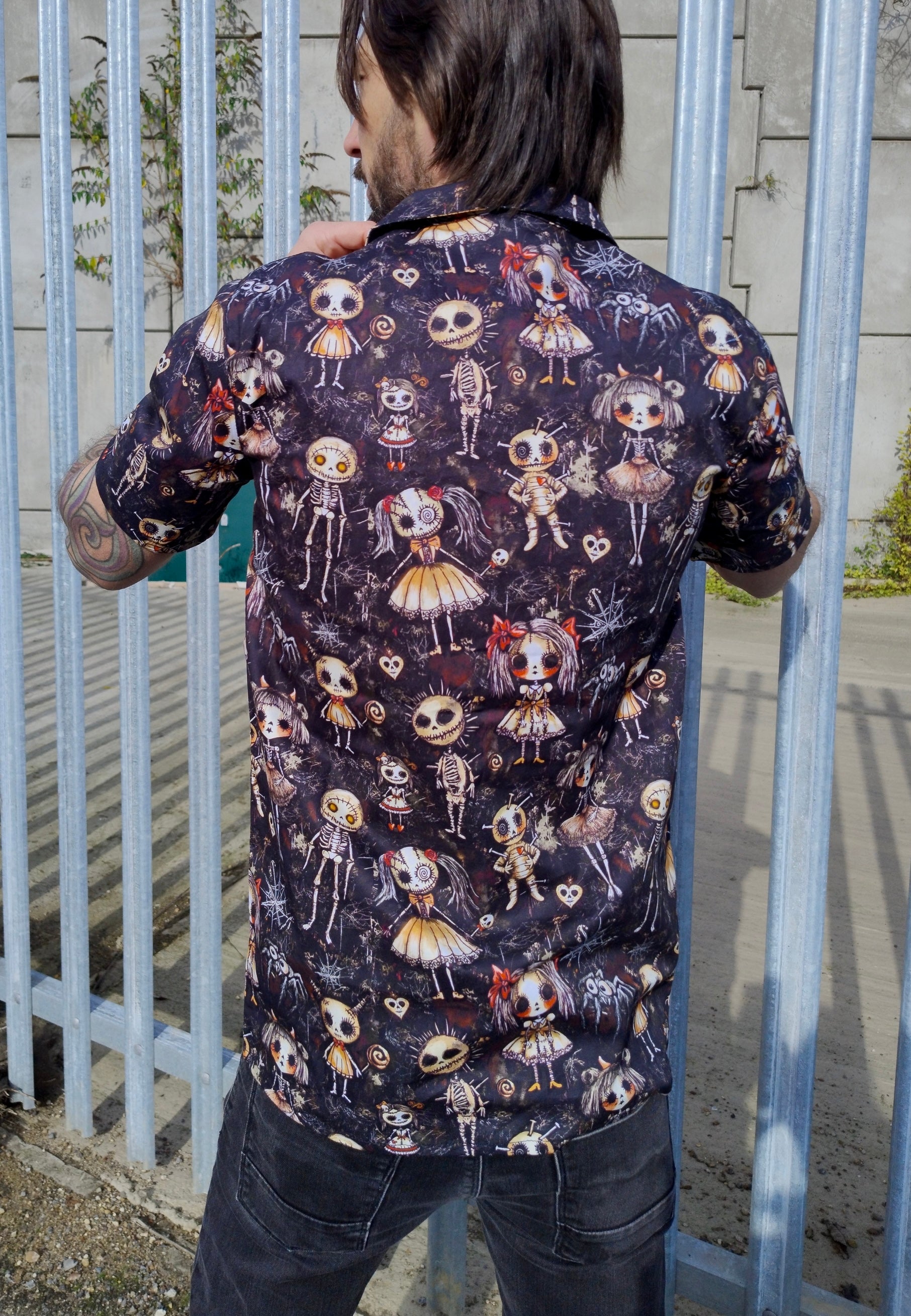 Back view of a dark haired man in a shirt with a Haunted Dolls print. Man is outdoors by an abandoned warehouse. 