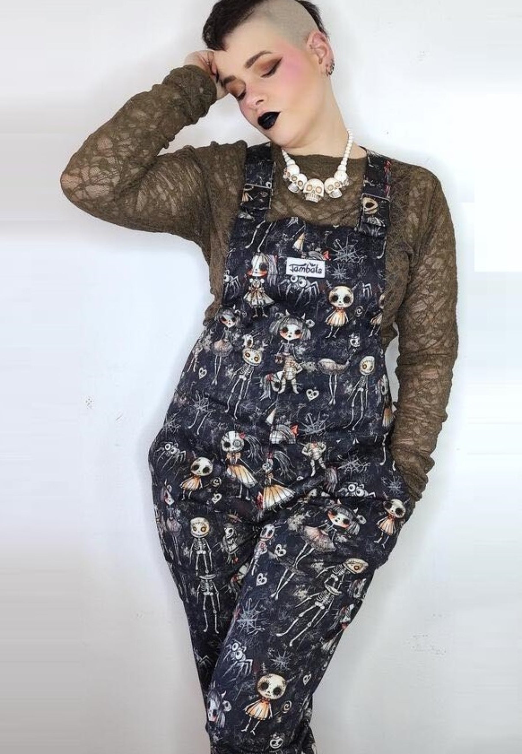 Model wearing dungarees with a pattern of cute but creepy haunted dolls in various poses against a dusty background. Model is also wearing a mesh top and skull necklace. Alternative Goth style. Jambats dungarees. 