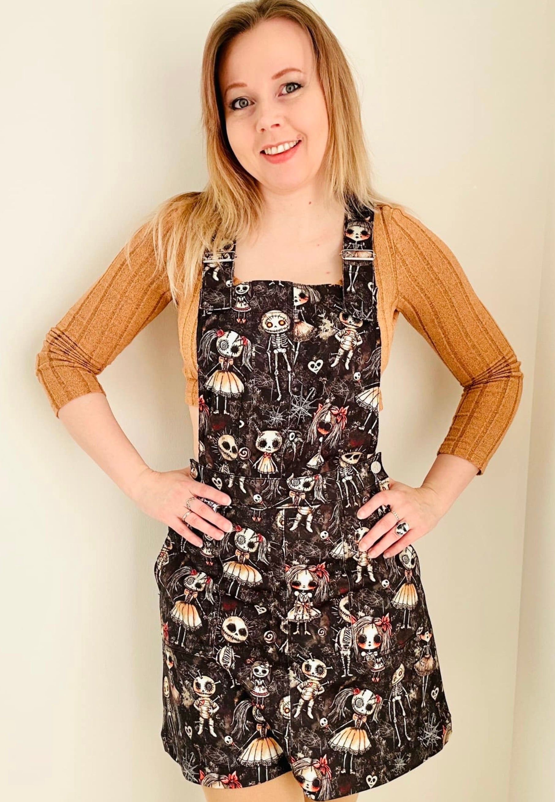 A woman wearing a stretch cotton twill pinafore dress with a haunted dolls print, featuring deep hip pockets and side button fastenings with unique bat buttons. The dolls print has cute but creepy dolls in a dusty, spiders web attic setting. 