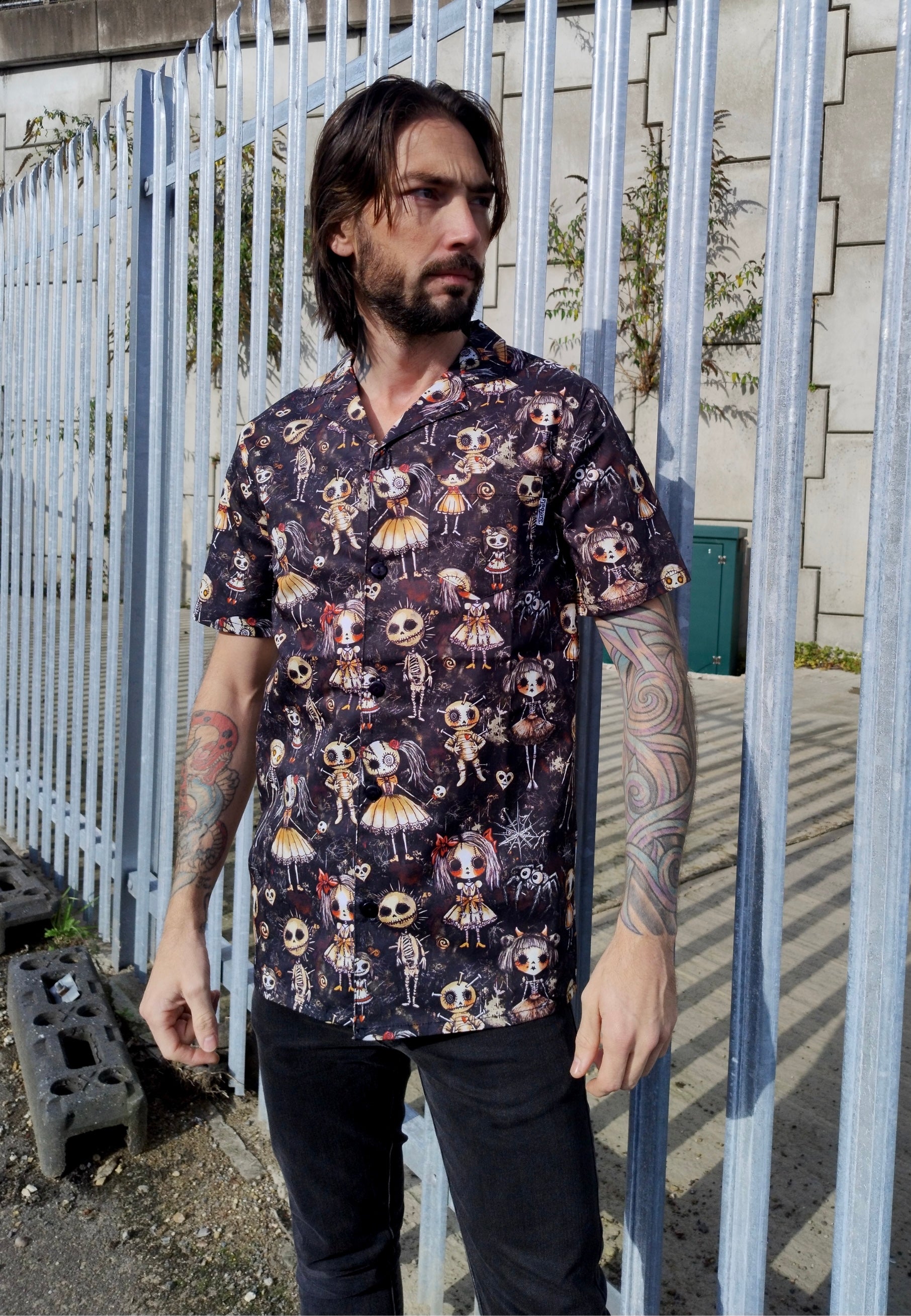 Man in a shirt with a Haunted Dolls print. Man is outdoors by an abandoned warehouse. 