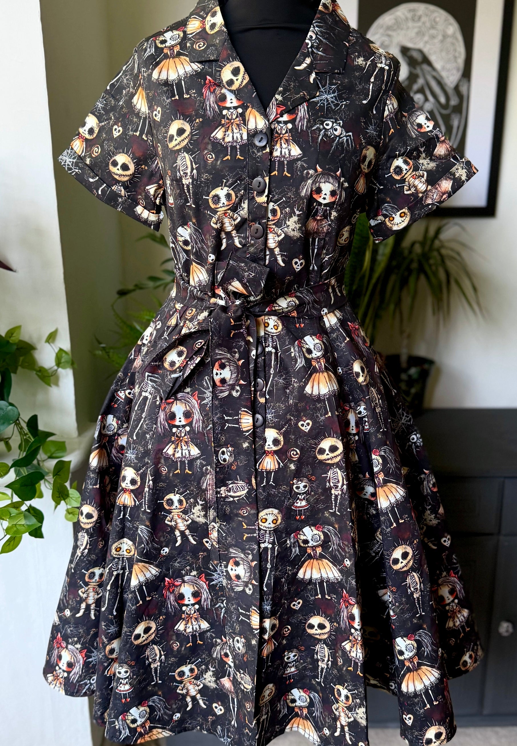 Beautiful shirt dress displayed on mannequin. Dress has a creepy haunted dolls print with a background of dusty cobwebs. 
