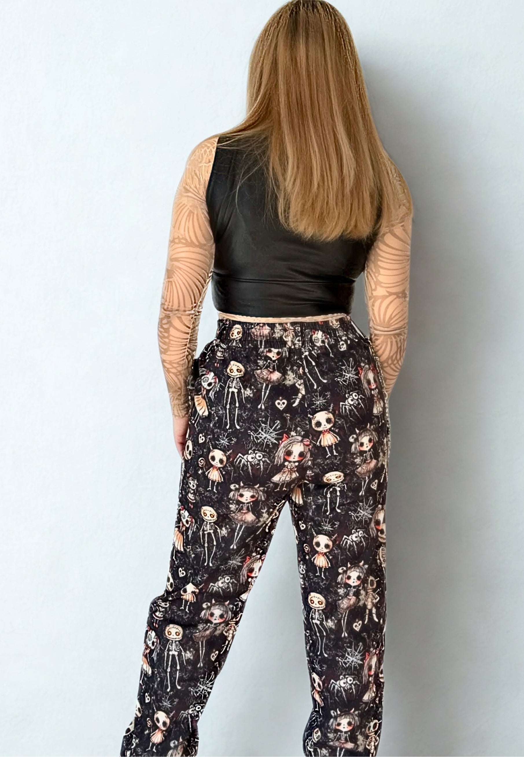Woman wearing a black leather top and gothic Haunted Dolls patterned trousers on a white studio background. Pictured from the back showing the back of the trousers 