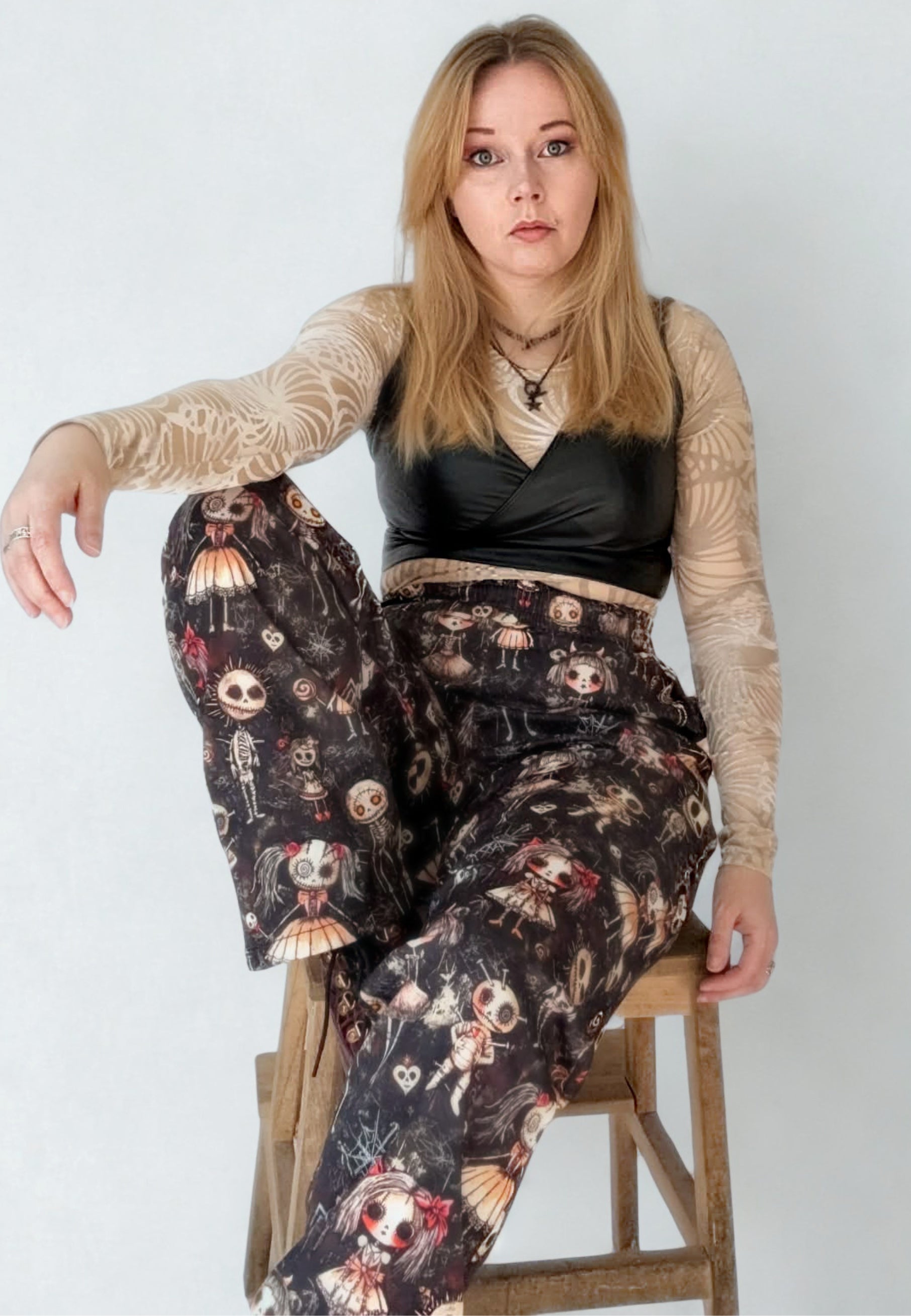 Woman wearing a black leather top and gothic Haunted Dolls patterned trousers on a white studio background. Woman is seated on a stool
