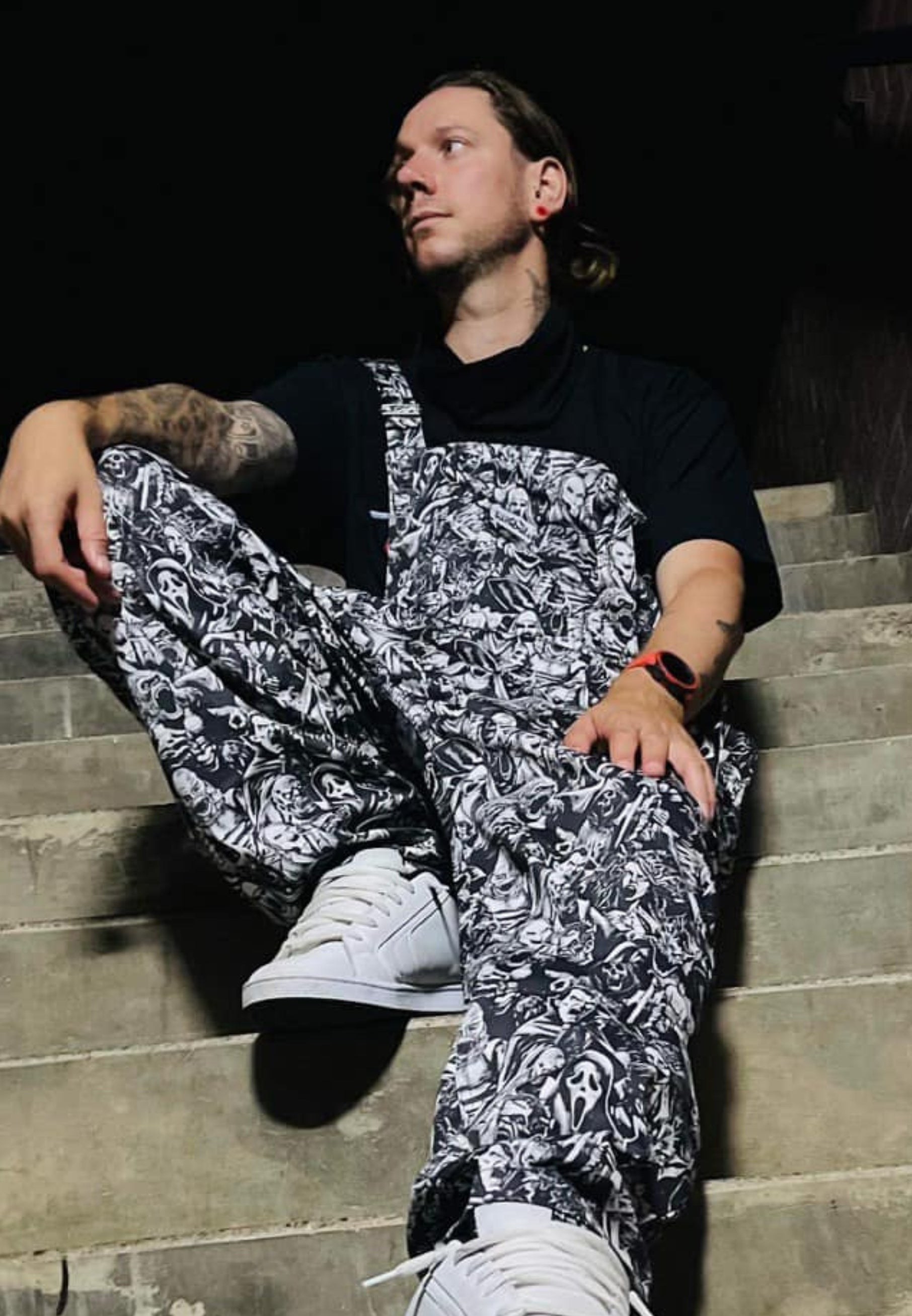 An alternative styled man  wearing black and white grayscale print dungarees over a black mesh top. Dungaree print is old school and modern monsters, horror characters and cryptids in an old horror comic style illustration. They are all screaming.