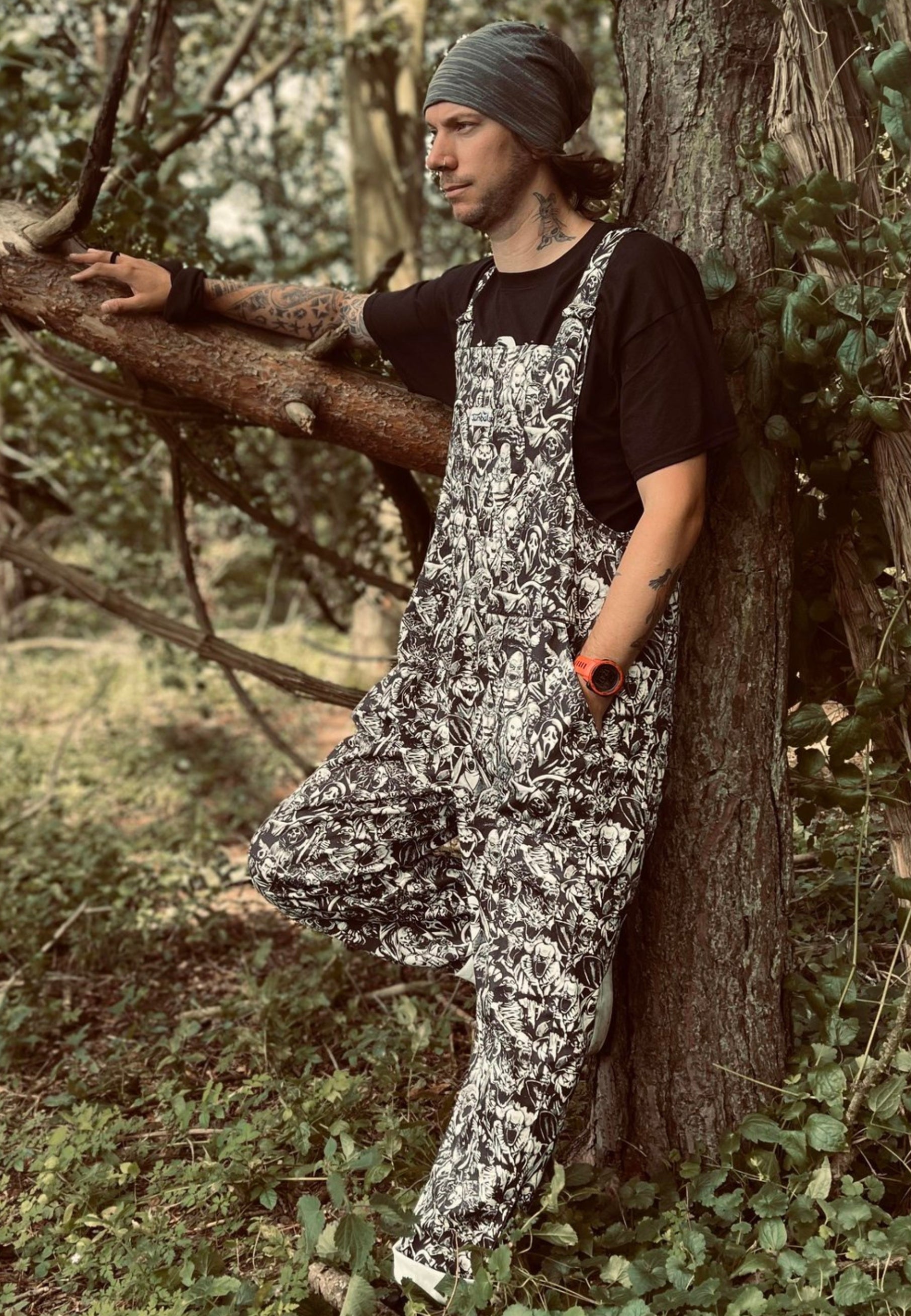 An alternative styled man outdoors standing by a tree  wearing black and white grayscale print dungarees over a black top. Dungaree print is old school and modern monsters, horror characters and cryptids in an old horror comic style illustration. They are all screaming.