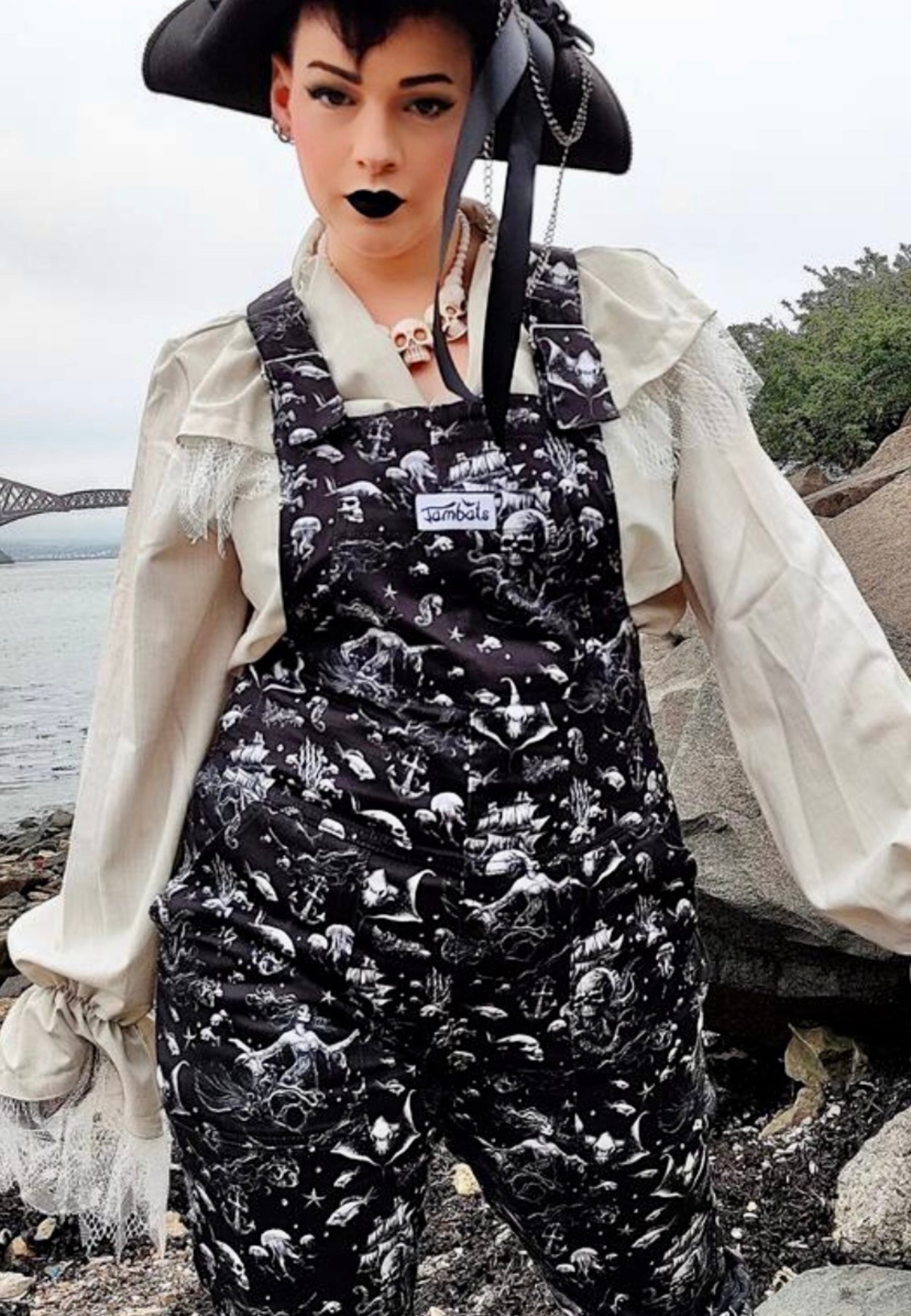 A woman wearing black dungarees with a pattern of ghostly pirate skull ships and eerie sirens. Styled with a white theatrical frilly shirt and black pirate hat.  Also skull necklace and gothic make up. 