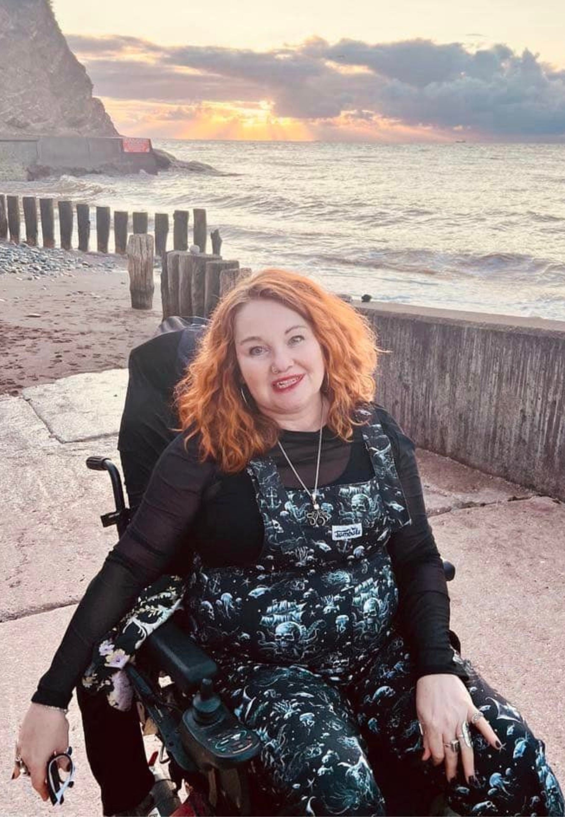 A woman wearing black dungarees with a pattern of ghostly pirate skull ships and eerie sirens. Styled with a black top. Woman is in a wheelchair in front of a beautiful sea view. 