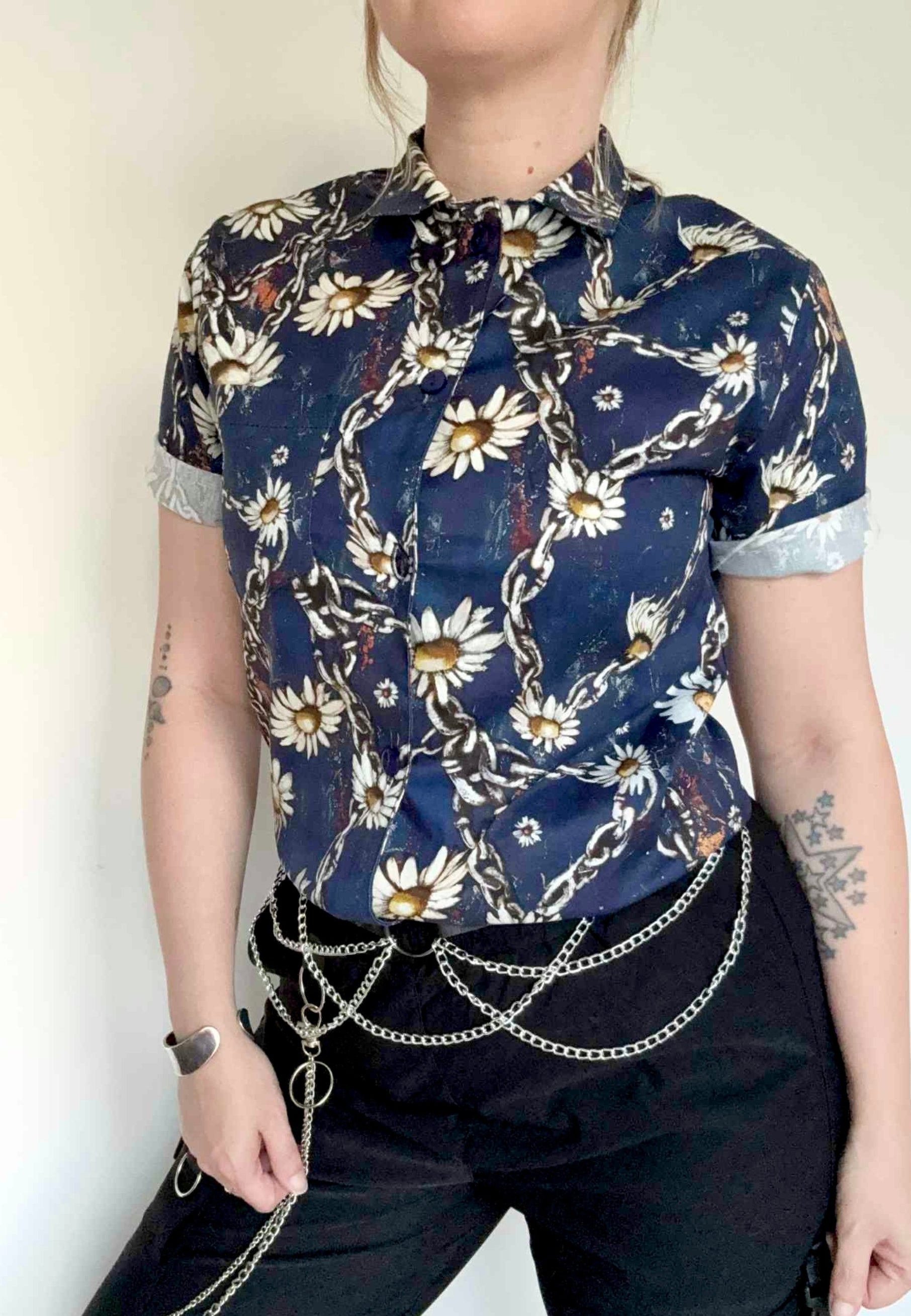 Woman wearing a deep blue shirt  with chains and daisies and dark pants against a plain background. Grungey floral style print. 