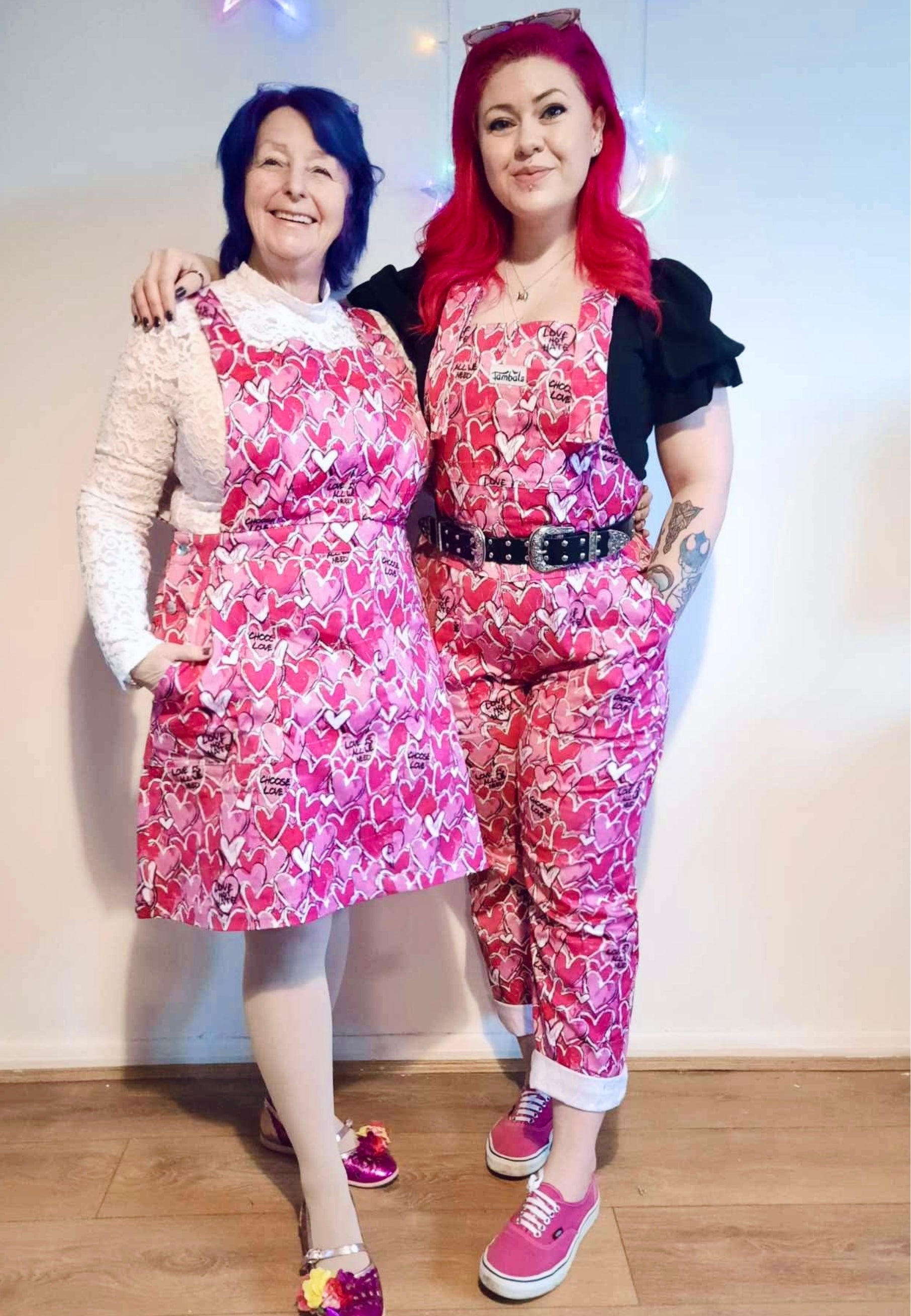Two women, one wearing a pink pinafore dress with a pattern of scribbled hearts and love related graphics and the other wearing the same print but in dungarees. 