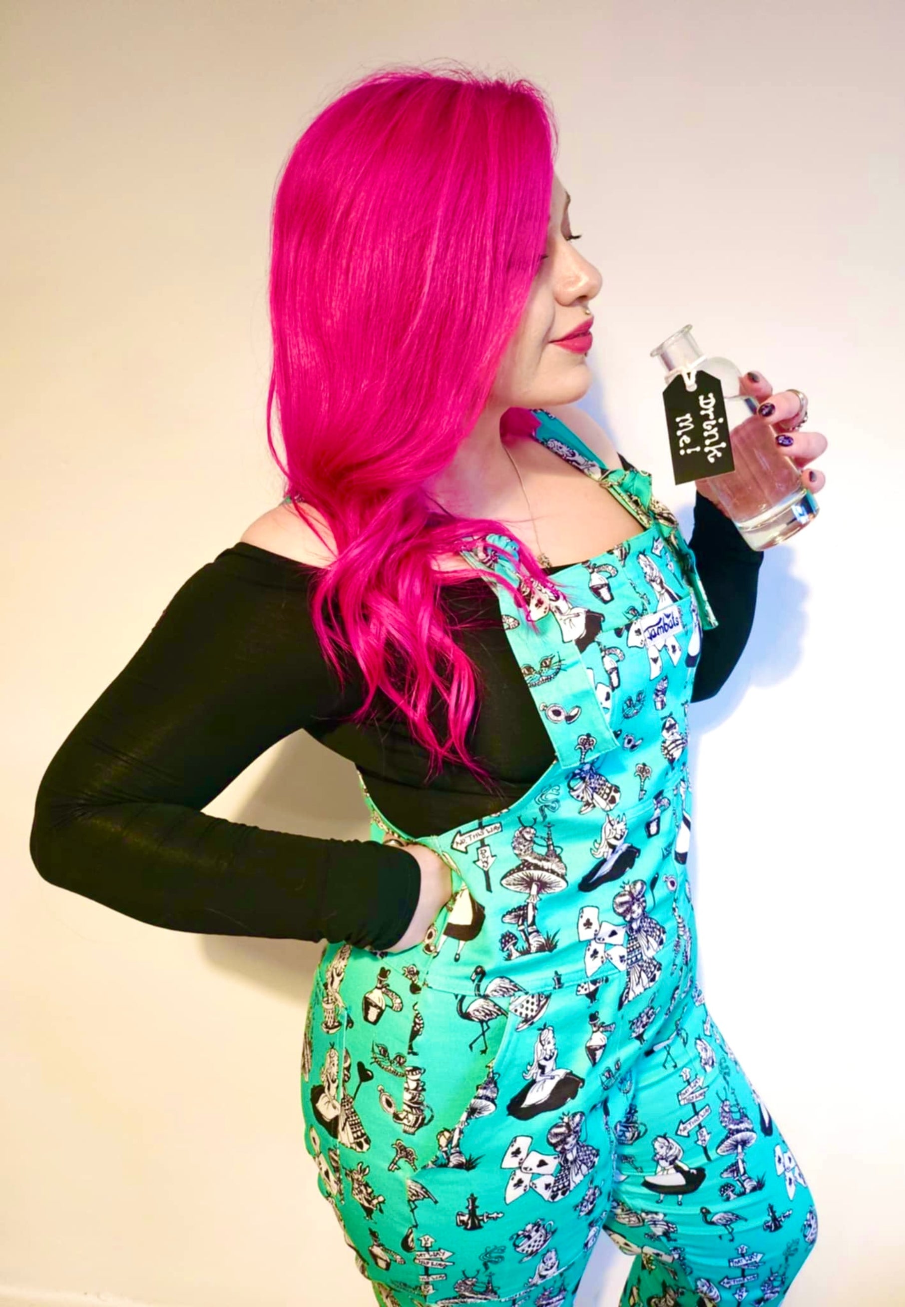 A woman with beautiful deep pink hair I’d wearing Alice in Wonderland print dungarees with black and white characters, standing outdoors.  woman is holding a ‘Drink Me’ bottle