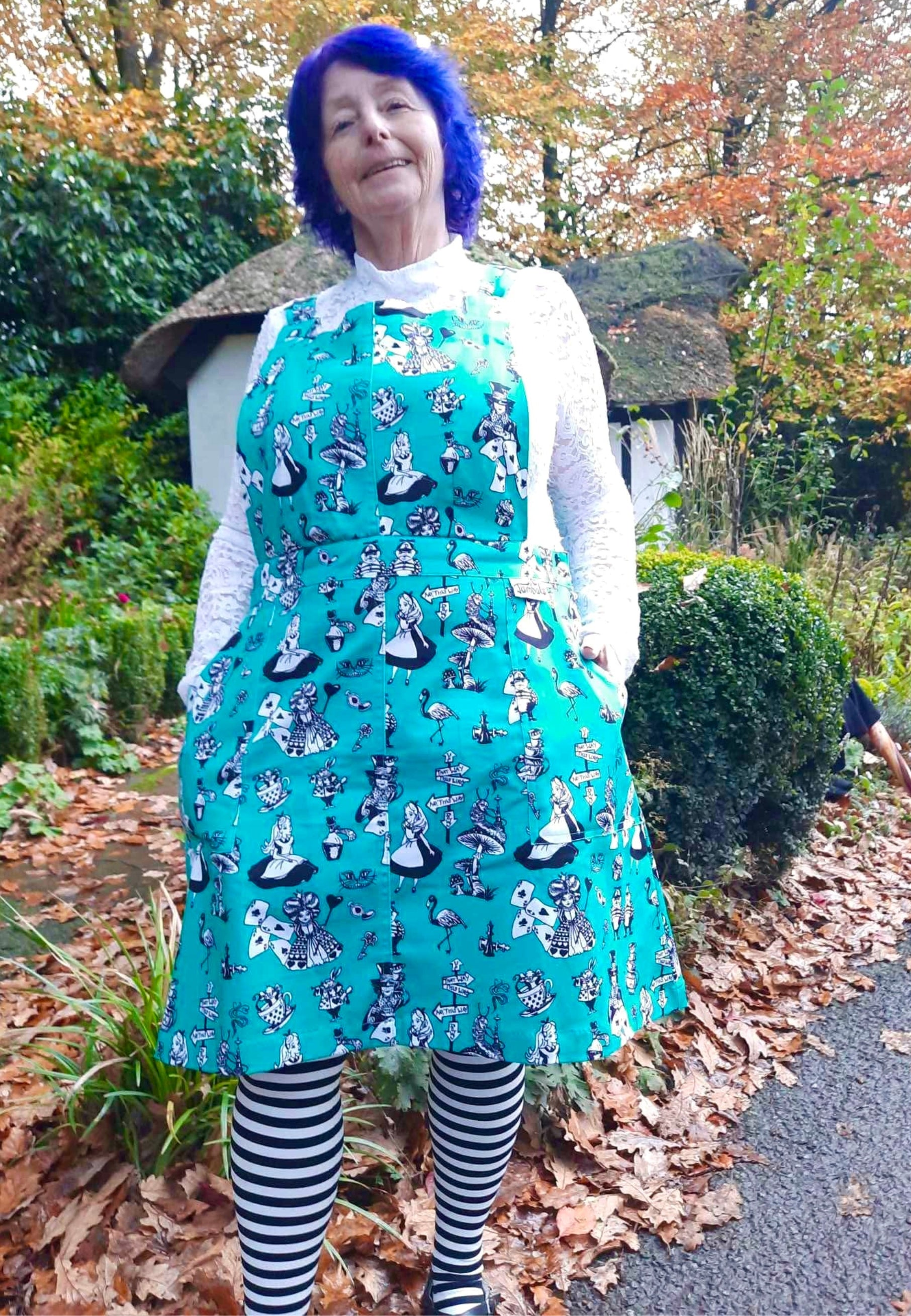 A woman wearing an Alice in Wonderland print pinafore dress with black and white characters, standing outdoors. Also wearing quirky black and white striped tights. 