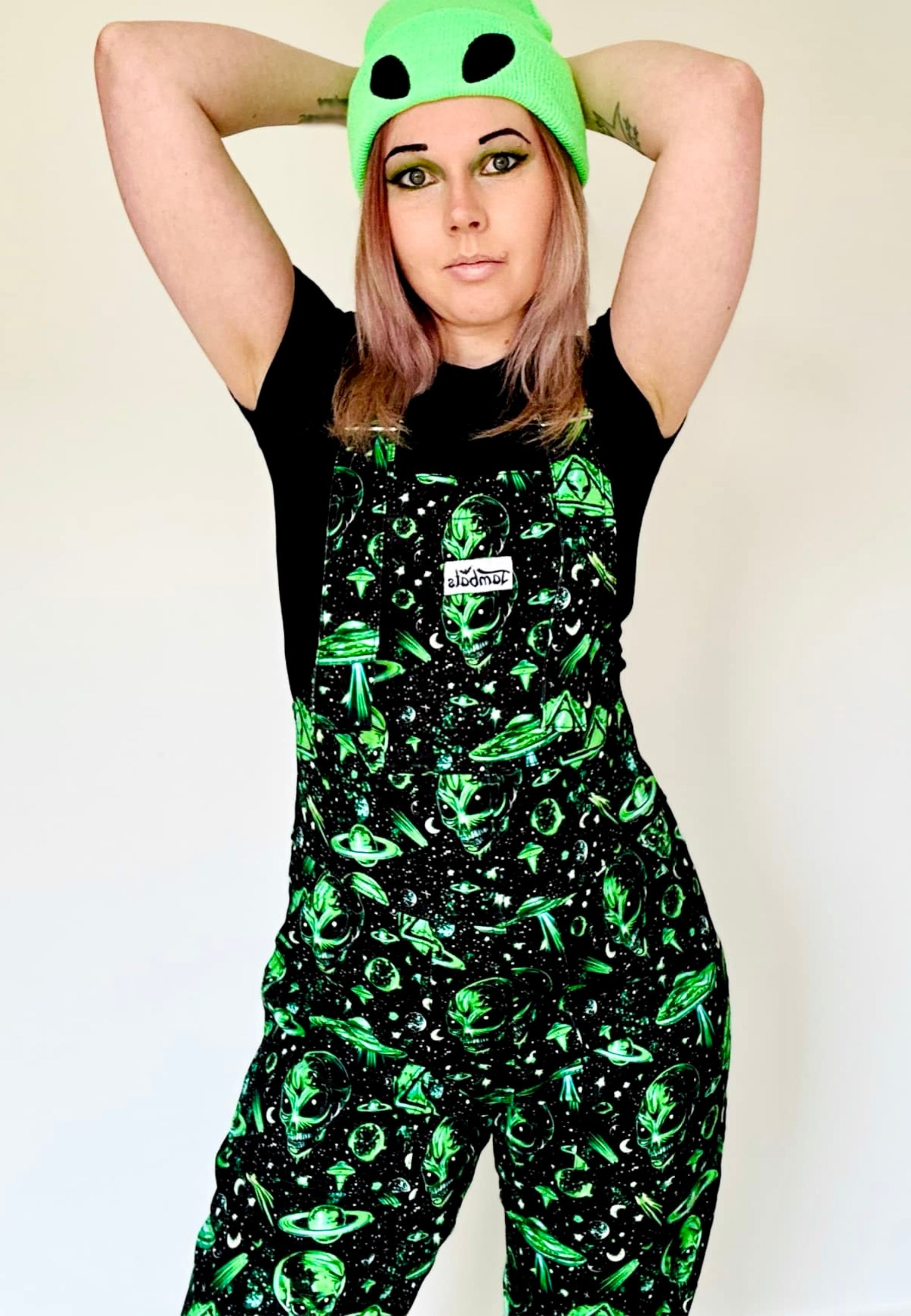 A woman wearing black and green dungarees with an extraterrestrial aliens print standing against a plain background and wearing a green alien beanie hat. 
