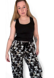 A woman modelling a pair of black and wide leg lounge pants with a beige and white alley cats print, paired with a black tank top, against a plain white background.