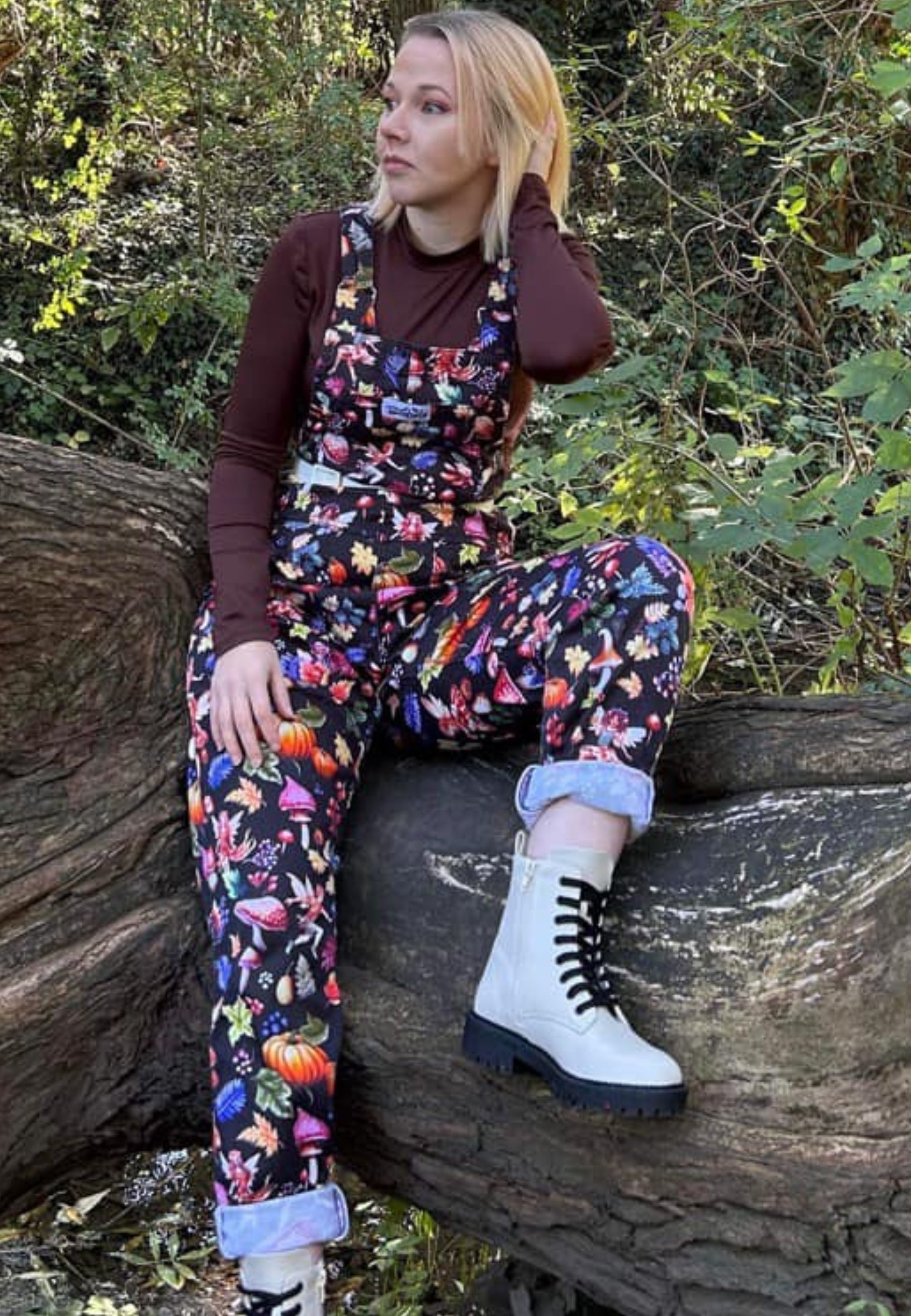 Woman sat on a tree log in the woods wearing dungarees in a magical Autumnal Fae print. Beautiful whimsical Fae dungarees
