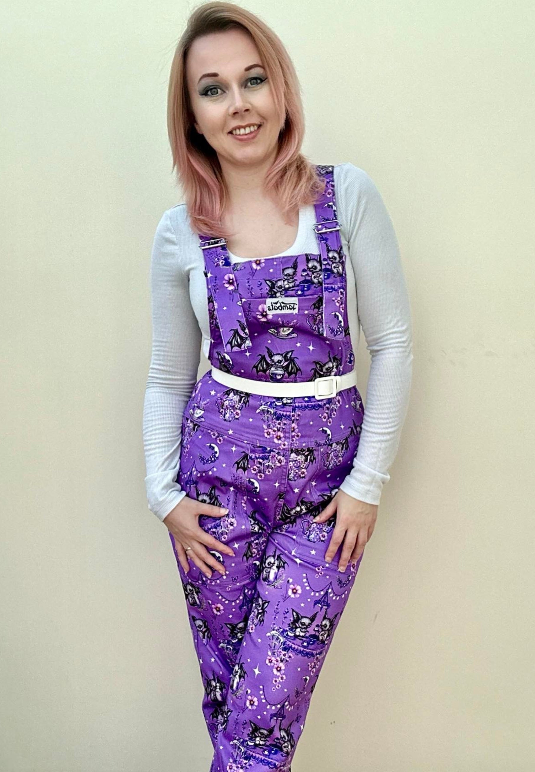 A woman wearing a deep lavender/soft purple dungarees with a cute gothic bats tea party print, paired with a white top and styled with a white belt.